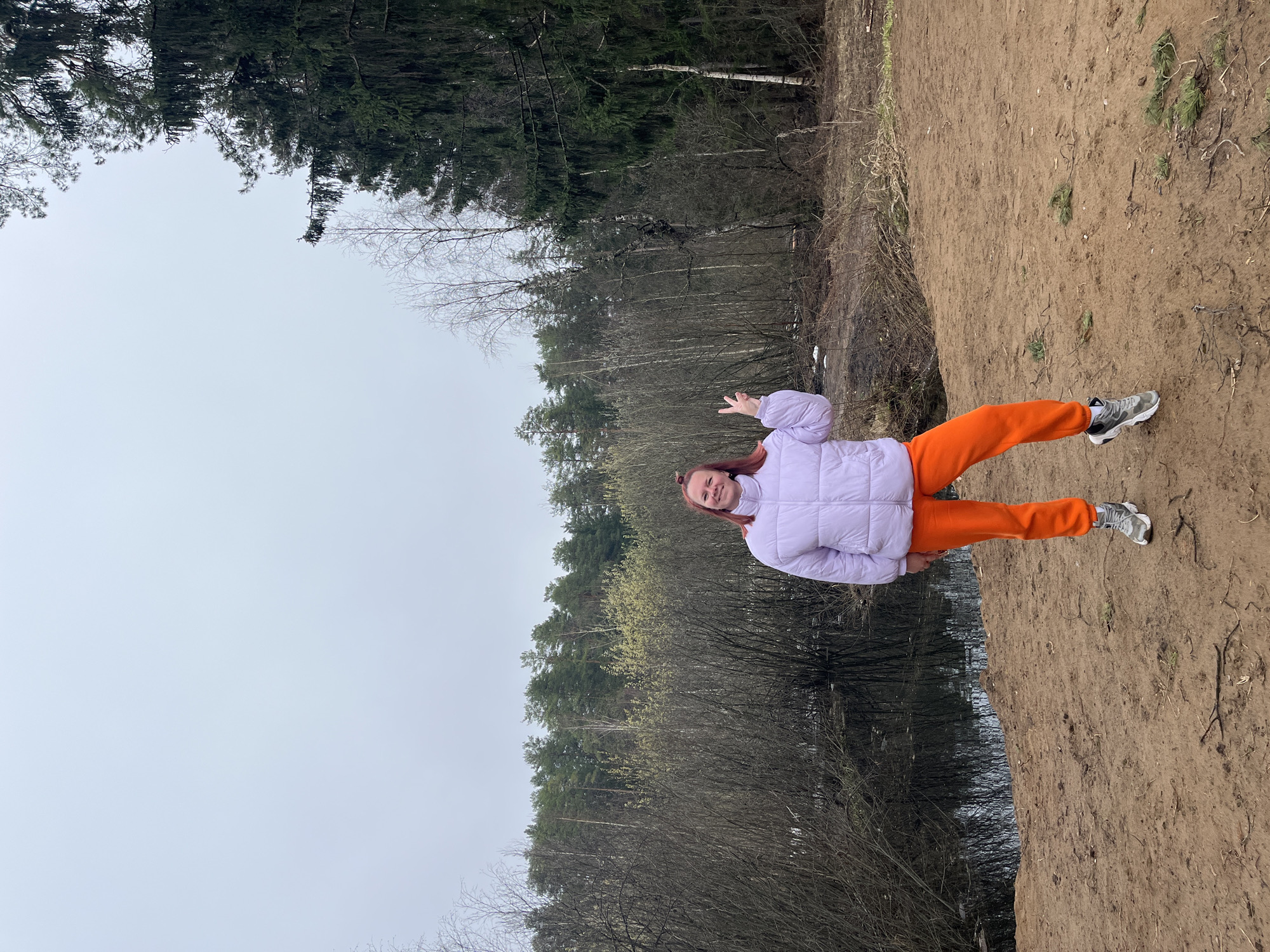 A young person wearing a light purple puffer jacket and bright orange sweatpants stands on a dirt embankment next to a body of water, potentially a river or lake, near a forest with both deciduous and evergreen trees. The person faces the viewer with a slight smile and a peace sign held up with their right hand. The sky is overcast. The location may be Chaikovsky, Russia, but this cannot be verified from the image alone.