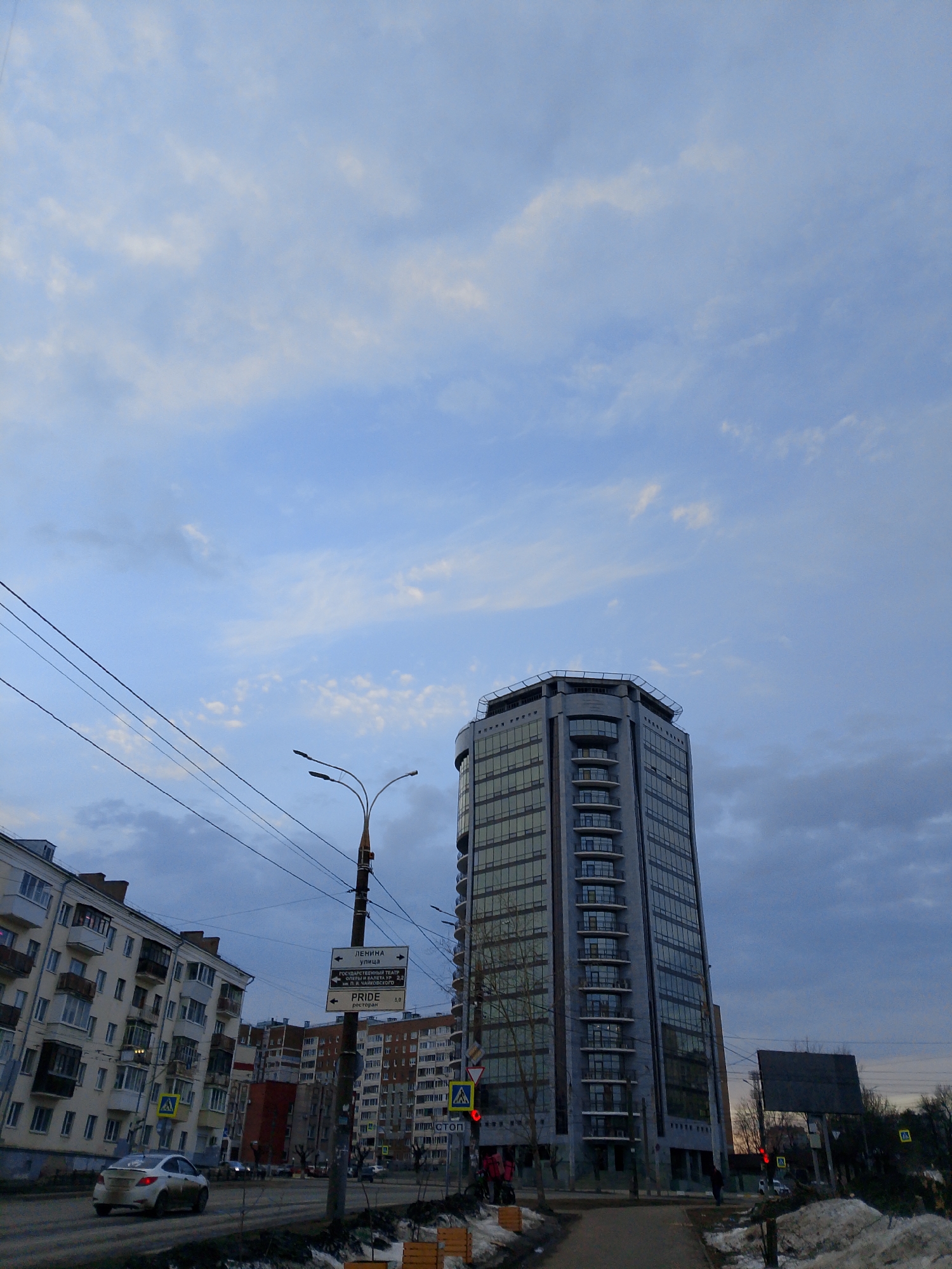 The image shows a street scene in Izhevsk, Russia, with a modern high-rise building dominating the view. The building is tall, gray, and has numerous windows, with a distinctive top section. In the foreground, there is a road with a car on it, as well as a sidewalk with patches of snow. Several older apartment buildings are visible in the background. Streetlights and power lines crisscross above the street. A street sign indicates directions, including "Lenina Street" and "PRIDE Restaurant". Traffic signals show red lights. The sky is overcast with blue and gray clouds. The overall impression is of a cold, urban environment in the late afternoon or early evening.