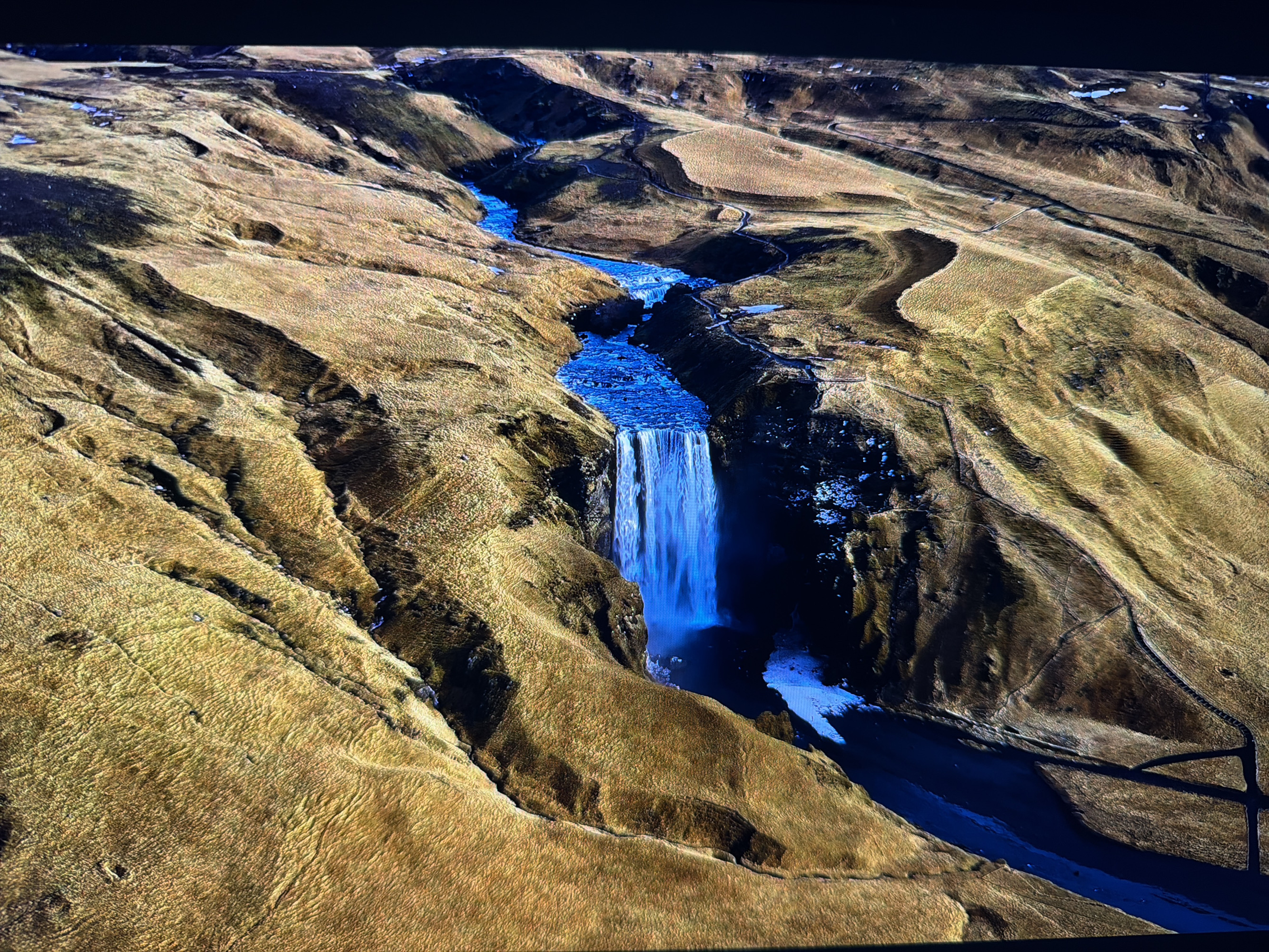 This image depicts an aerial view of a waterfall cascading down a rocky cliff face into a river. The surrounding landscape is characterized by rugged, brown, and dry vegetation, with some patches of snow or ice visible on the ground. The river flows through a canyon, with winding paths visible along its edges. The overall scene is natural and remote, with no immediate signs of urban development. There are no people or discernible man-made structures in the immediate vicinity of the waterfall or the canyon. The lighting suggests it might be daytime, with shadows indicating variations in terrain. The image does not contain any identifiable landmarks or features that would place it specifically in the city of Kafr Yasif, Israel.