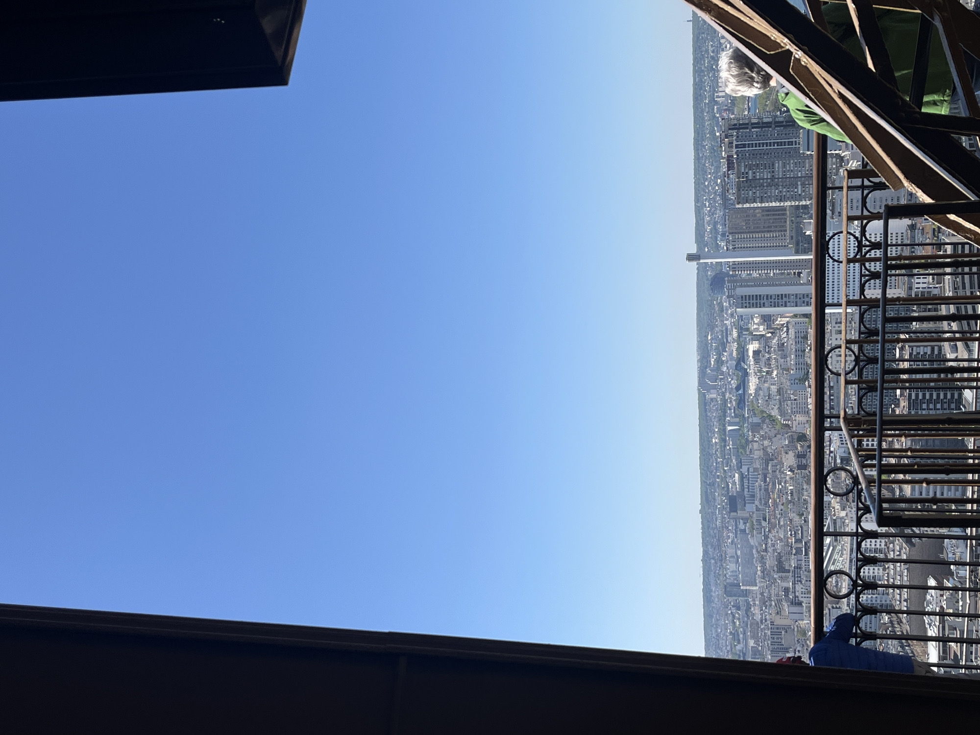 View from the Eiffel Tower in Paris, France. The cityscape is visible beyond a metal railing. The Tour Montparnasse skyscraper is prominent in the distance. People are visible, including one with gray hair and a green jacket. The framing of the shot includes the structure of the Eiffel Tower. The sky is blue.