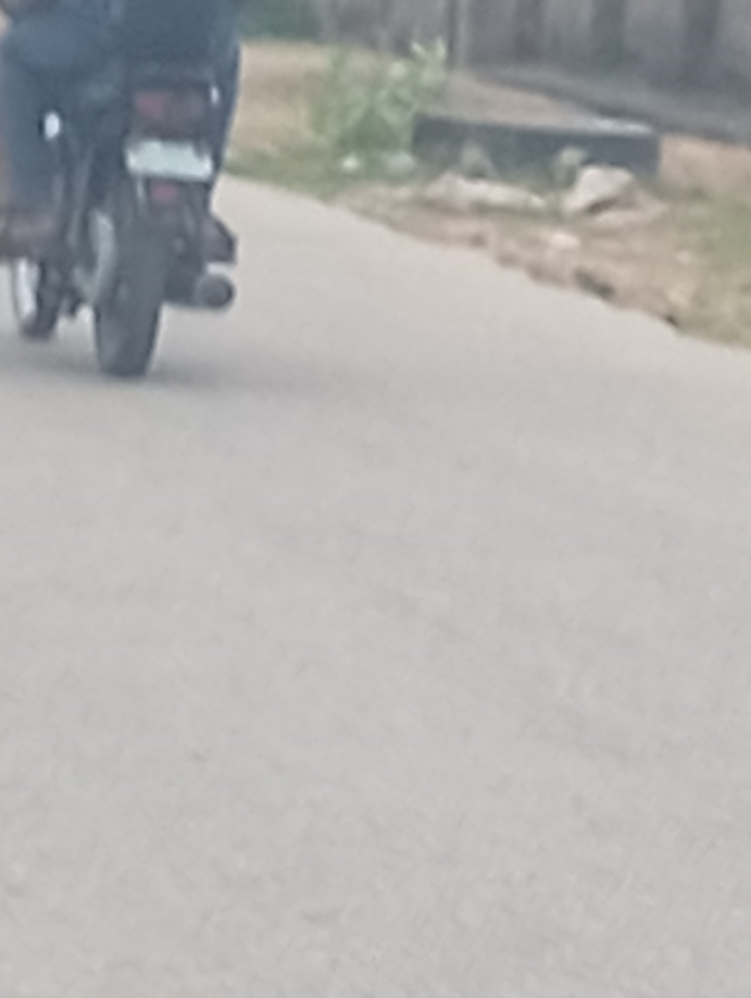 The image shows a rear view of a motorcycle with a rider from the city of Adankolo, Nigeria. The motorcycle is on a paved road, and the rider is wearing dark pants. The rear of the motorcycle features a license plate, a taillight, and an exhaust pipe. In the background, there is some vegetation and what appears to be a tire and rocks near the side of the road. The scene is slightly blurry, suggesting the photo may have been taken while in motion.