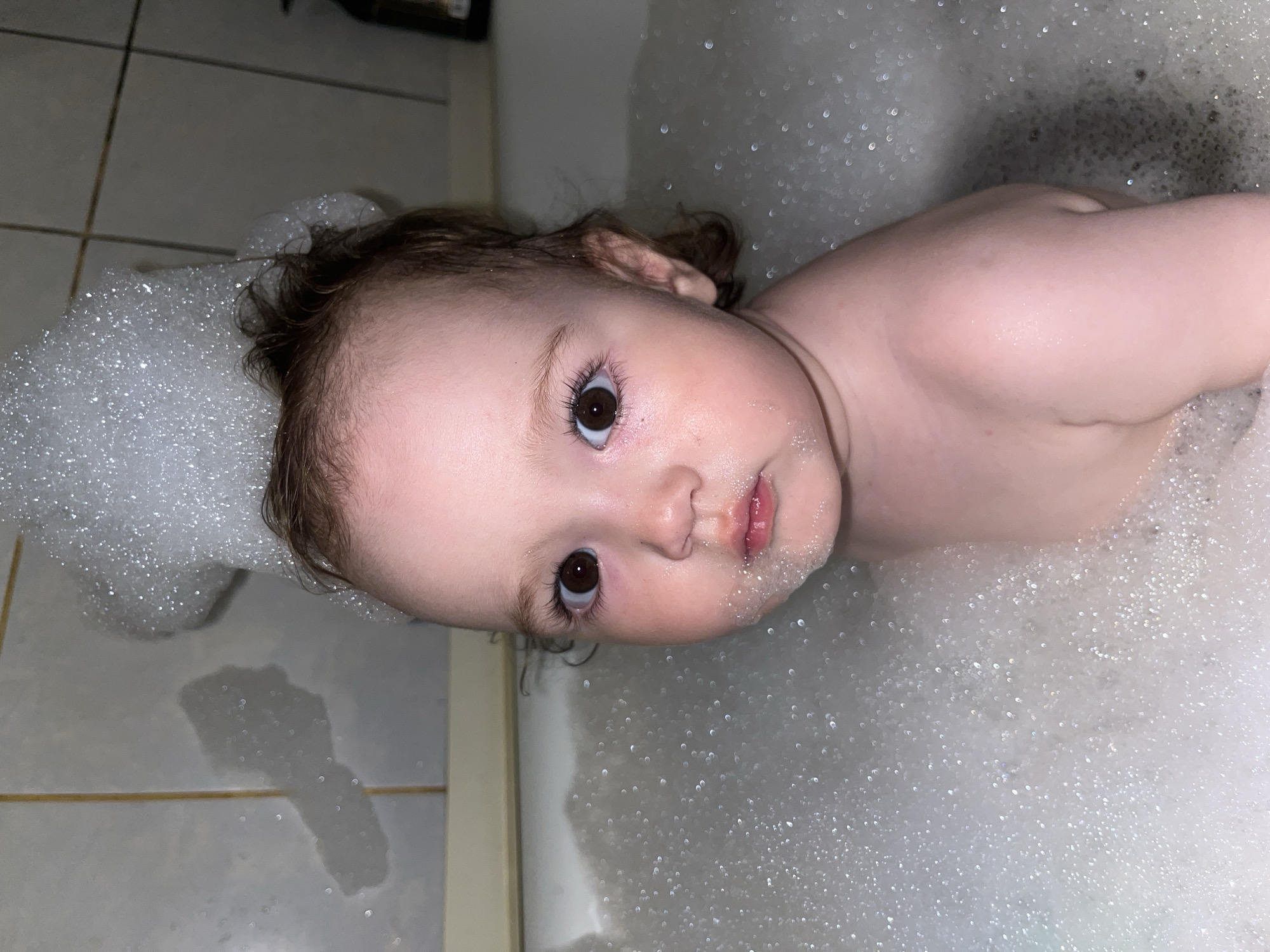 A baby is lying in a bathtub filled with bubbles. The baby has brown hair and brown eyes. The bathtub is in a bathroom with tile walls. The location is not evident in the image.