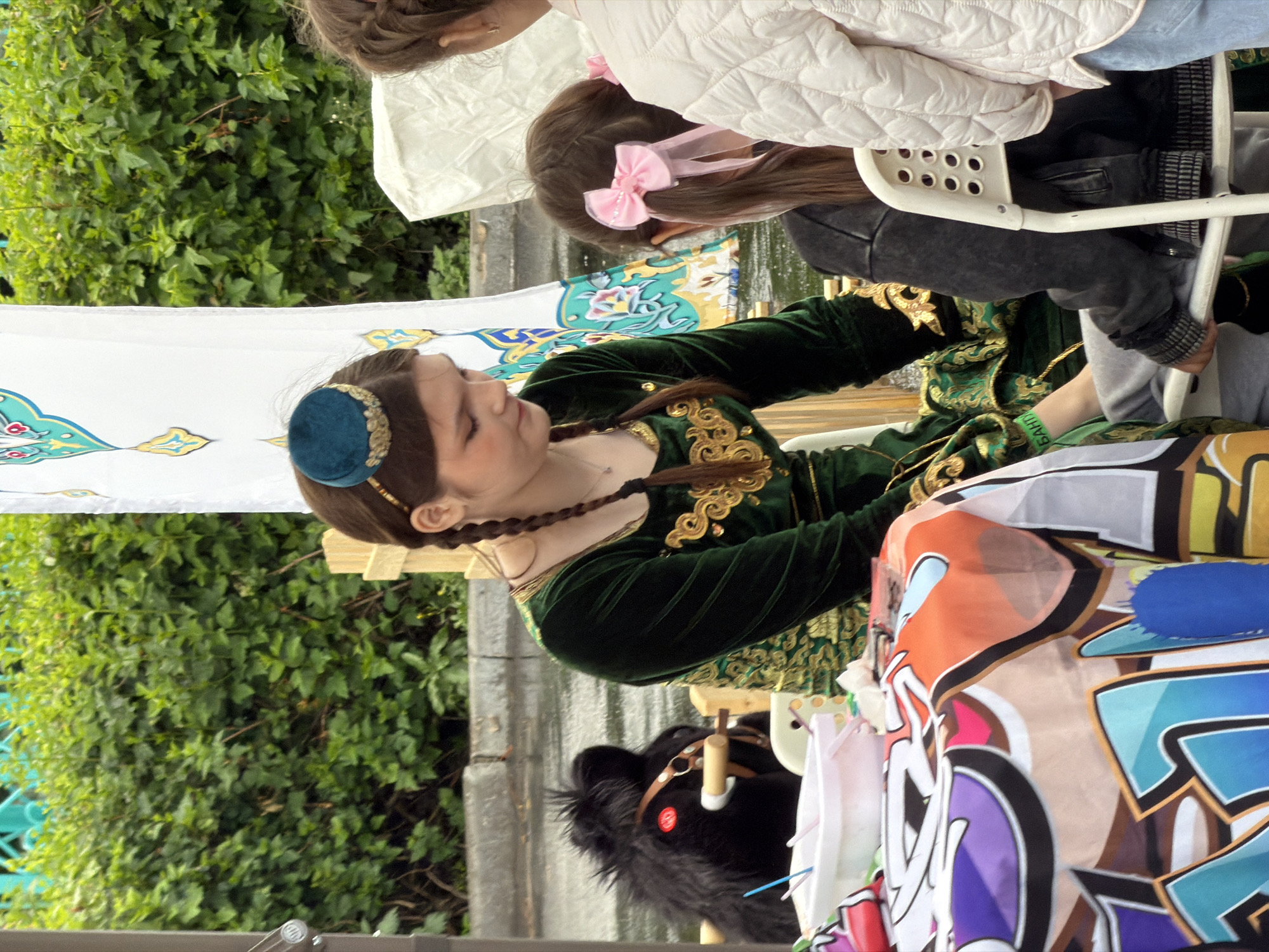 In an outdoor setting with lush green foliage in the background, a young woman in a green velvet dress adorned with gold embroidery and a small blue hat sits at a table covered with a colorful cloth. She is looking down, possibly at something on the table, and her hair is styled in two braids. To her left, a black stuffed toy horse with a red tag is visible. In front of the woman, a white bowl with colorful straws is on the table. To the woman's right, a young girl with her back to the camera, wearing a black jacket and pink hair bows, sits on a white chair. Another person, whose back is partially visible, stands to the right, wearing a quilted off-white jacket. The ground appears wet, suggesting recent rain.