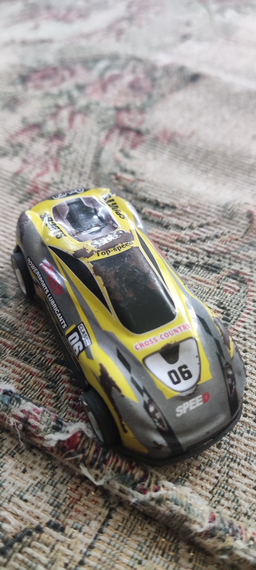 A close-up shot shows a toy racing car with a yellow and gray paint scheme. The car has the words "Top-speed," "Cross Country," and "Speed" visible on its body. The number "06" is prominently displayed on the front of the car. The toy appears slightly worn, with some scuff marks and faded paint. The car is positioned on a textured surface, possibly a rug or blanket, with a floral pattern in muted colors. The image does not provide any information about the location in Gebang, Indonesia, as there are no identifiable landmarks or environmental cues to suggest such a location.