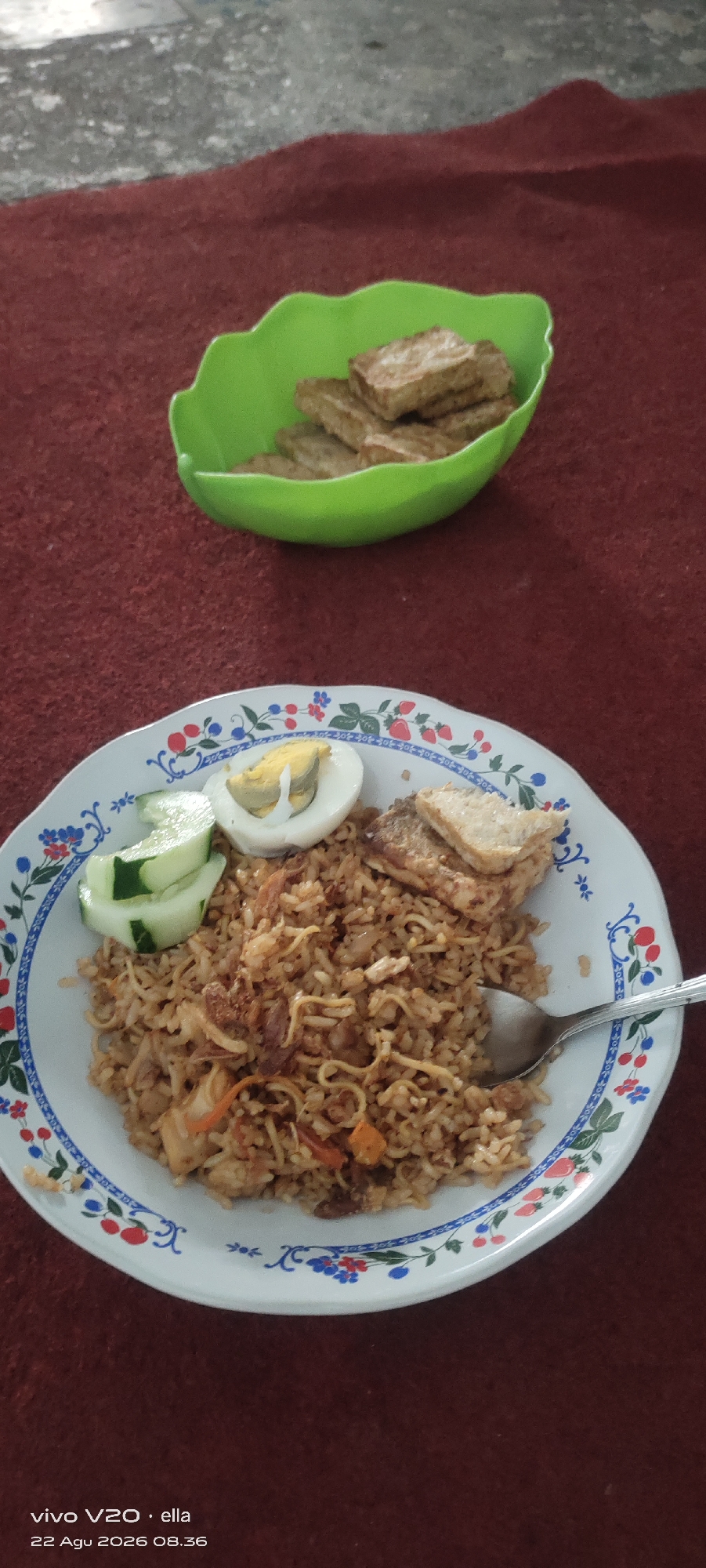 A close-up, high-angle shot shows a plate of fried rice with side dishes on a maroon carpet. The plate is white with a decorative blue and red floral rim. The fried rice contains noodles and what appear to be pieces of meat and vegetables. Garnishings on the plate include two slices of cucumber, a hard-boiled egg cut in half, and two fried pieces of tofu or tempeh. To the upper right of the plate, in a green plastic bowl, are more pieces of fried tofu or tempeh. The background is a blurred maroon carpet and a greyish concrete surface. The bottom left corner of the image displays a timestamp: "vivo V20 · ella 22 Agu 2026 08.36". The scene appears to be in a residential setting, possibly a home in Simo, Indonesia, given the timestamp's date format and language. There are no people visible in the image.
