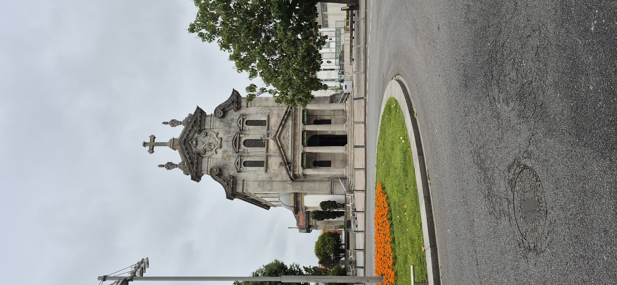 The image shows the Igreja de Nossa Senhora da Conceição, a church located in Póvoa de Varzim, Portugal. The church has a neoclassical facade with a large stone cross at the top. In front of the church, there is a circular garden with orange flowers and green grass, bordered by a gray concrete curb. A paved road with a manhole cover in the foreground curves around the garden. A street lamp is visible to the left of the church. A tree with green foliage stands to the right of the church. The sky is overcast. There are no people visible in the image.
