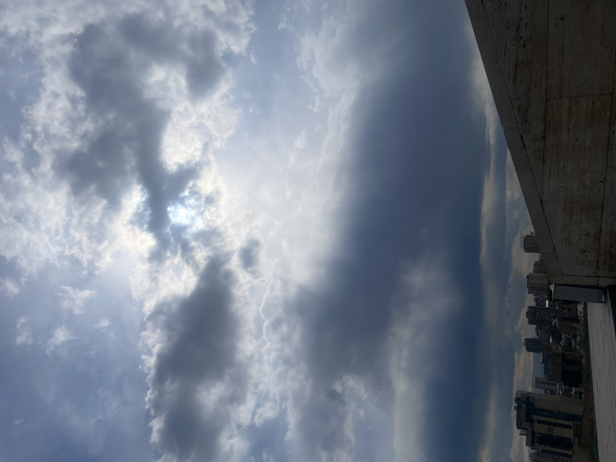 The image depicts a cityscape view from a high vantage point, likely a building in Yerevan, Armenia. To the left, the sky is filled with mostly grey and some white clouds, partially obscuring the sun. Light rays appear to stream through gaps in the clouds. A building structure, possibly a rooftop or balcony, is visible along the right edge of the frame. Below the cloud line, a cluster of buildings and structures, characteristic of an urban setting, extends towards the bottom of the frame. The sky is casting some light on buildings, while it also appears that weather is overcast.