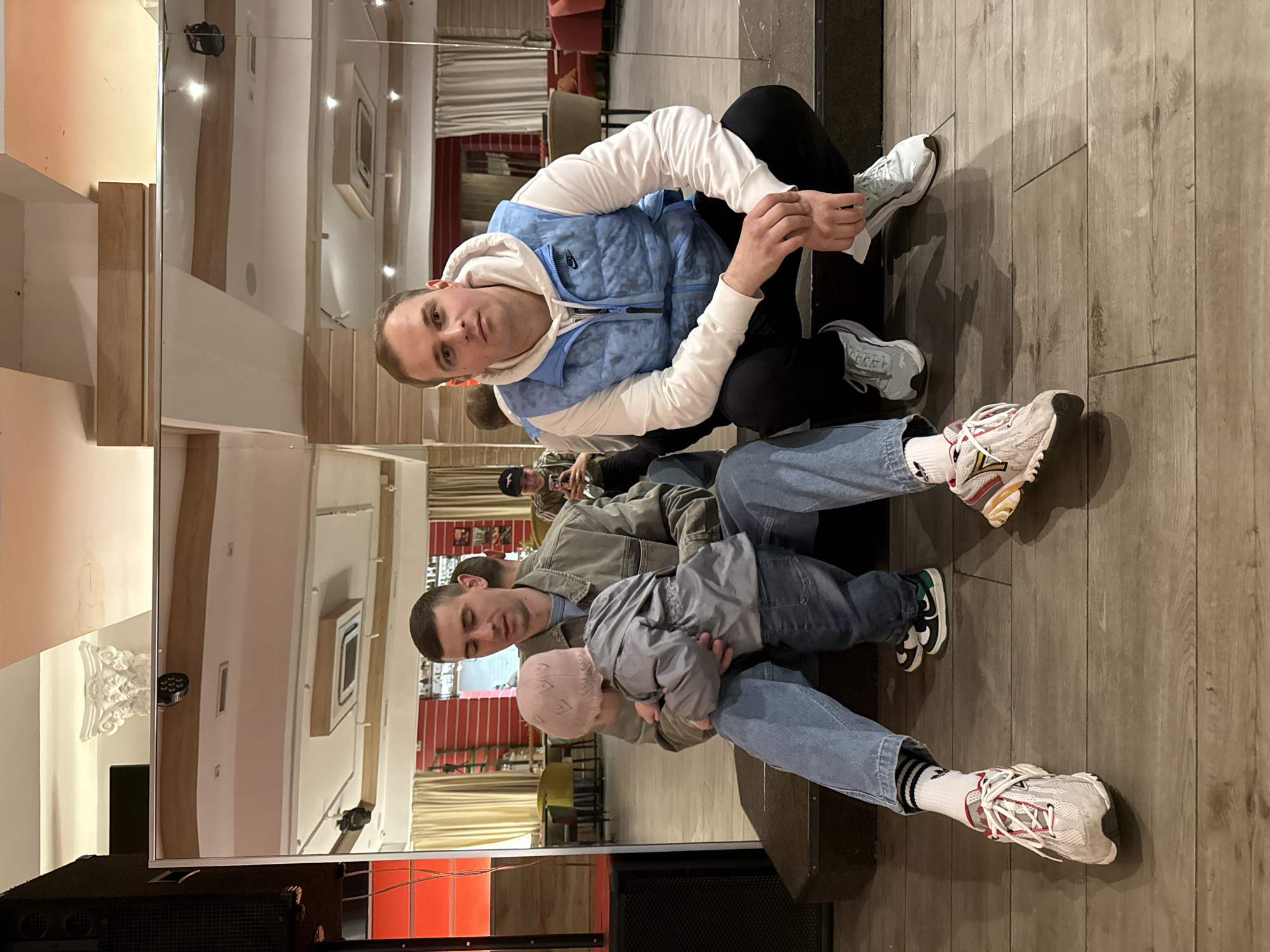 Two men are sitting on a low platform in front of a large mirror that reflects the interior of a room. The man in the foreground wears a light blue jacket over a white hooded sweatshirt and black pants and white shoes. The man is looking at the camera. The man in the background wears a tan jacket and blue jeans and white socks, and is holding a child wearing a pink cap and gray jacket. Both men are looking at the camera. The setting appears to be indoors with a wooden floor and a mirror that reflects a dining area with tables and chairs. Based on architectural styles and the appearance of the interior décor, the location is a restaurant or event space in Moscow, Russia.