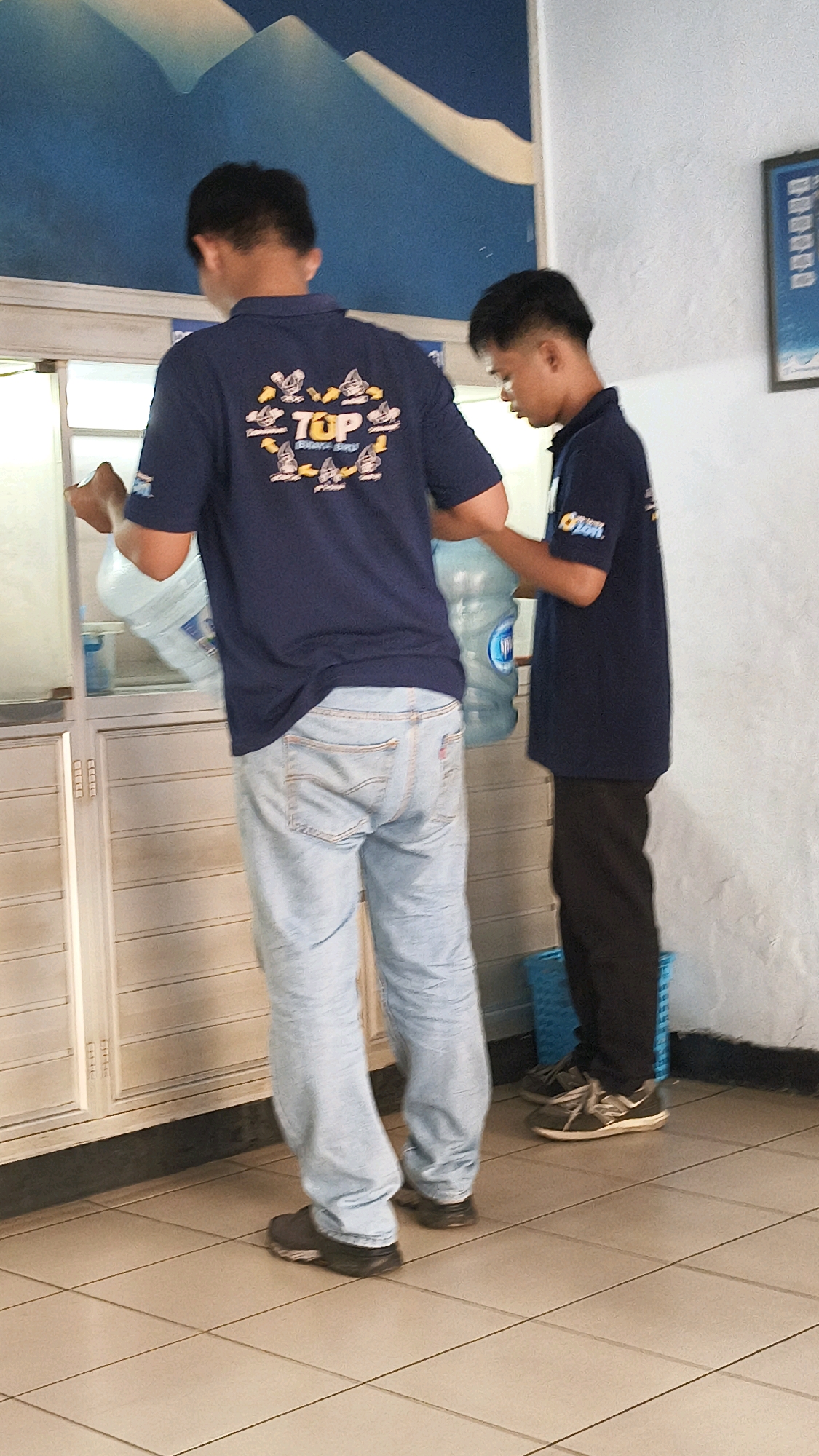 Two young men are standing in front of a counter, possibly in a service station in Bekasi, Indonesia. The man on the left is wearing a dark blue t-shirt with a graphic design on the back and light blue jeans. He is holding a large, clear plastic water jug with a blue handle. The man on the right is wearing a dark blue t-shirt and black pants. He is also holding a large, clear plastic water jug. The environment appears to be indoors, with tiled flooring and a wall painted white or a light color. A blue plastic basket is visible on the floor near the man on the right. The lighting suggests it is daytime, and the overall scene is casual.