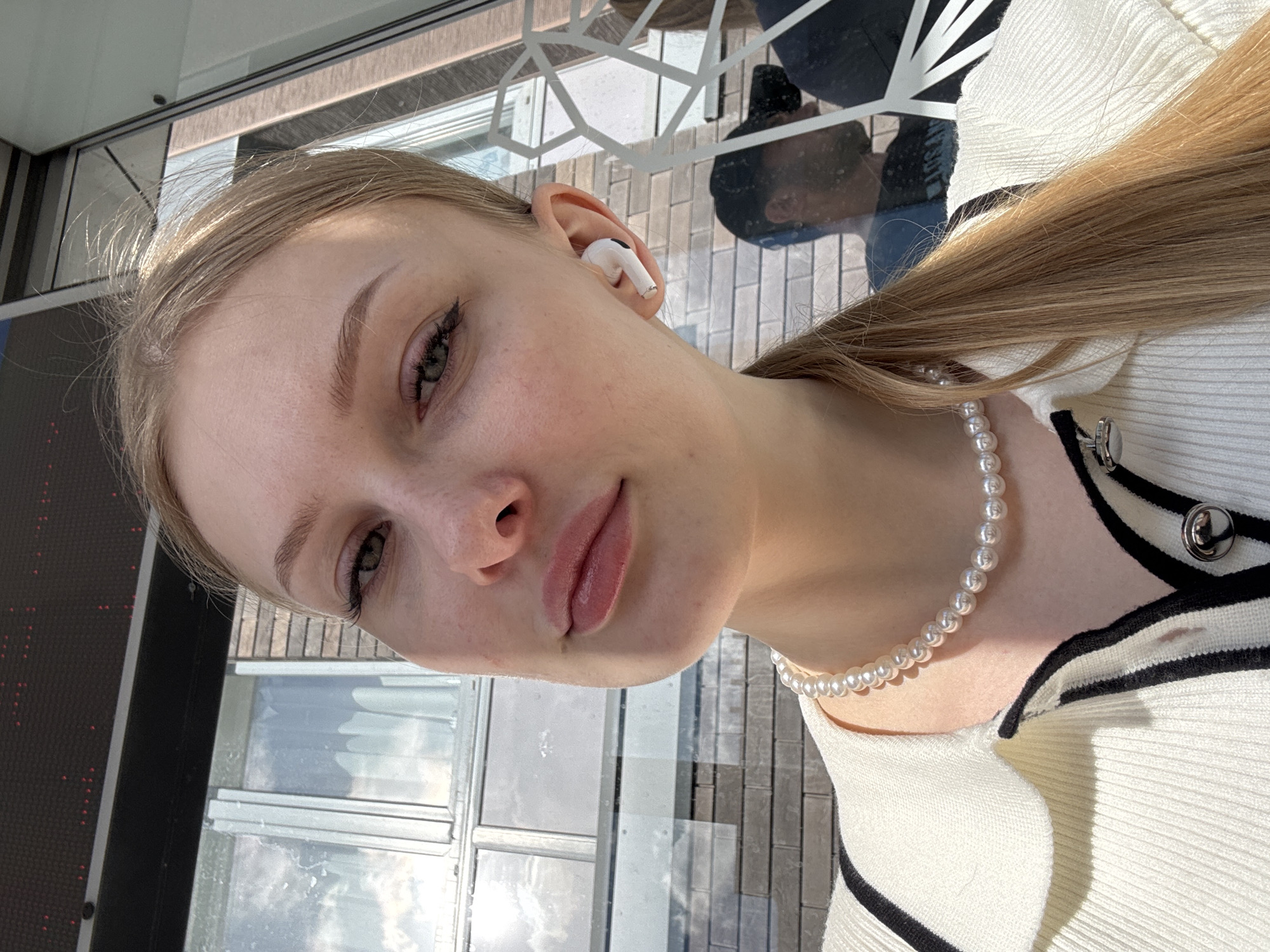 A young woman with blonde hair is taking a selfie in an outdoor urban setting, possibly a street or a building facade with reflective surfaces. She is wearing a white collared shirt with black trim and a pearl necklace. She has on white earbuds. The background shows a brick wall and windows, with reflections of people and the sky. The lighting suggests it is daytime. The location is not specified in the image, but if it is in Perm, Russia, it could be any city street or public area with similar architectural elements.