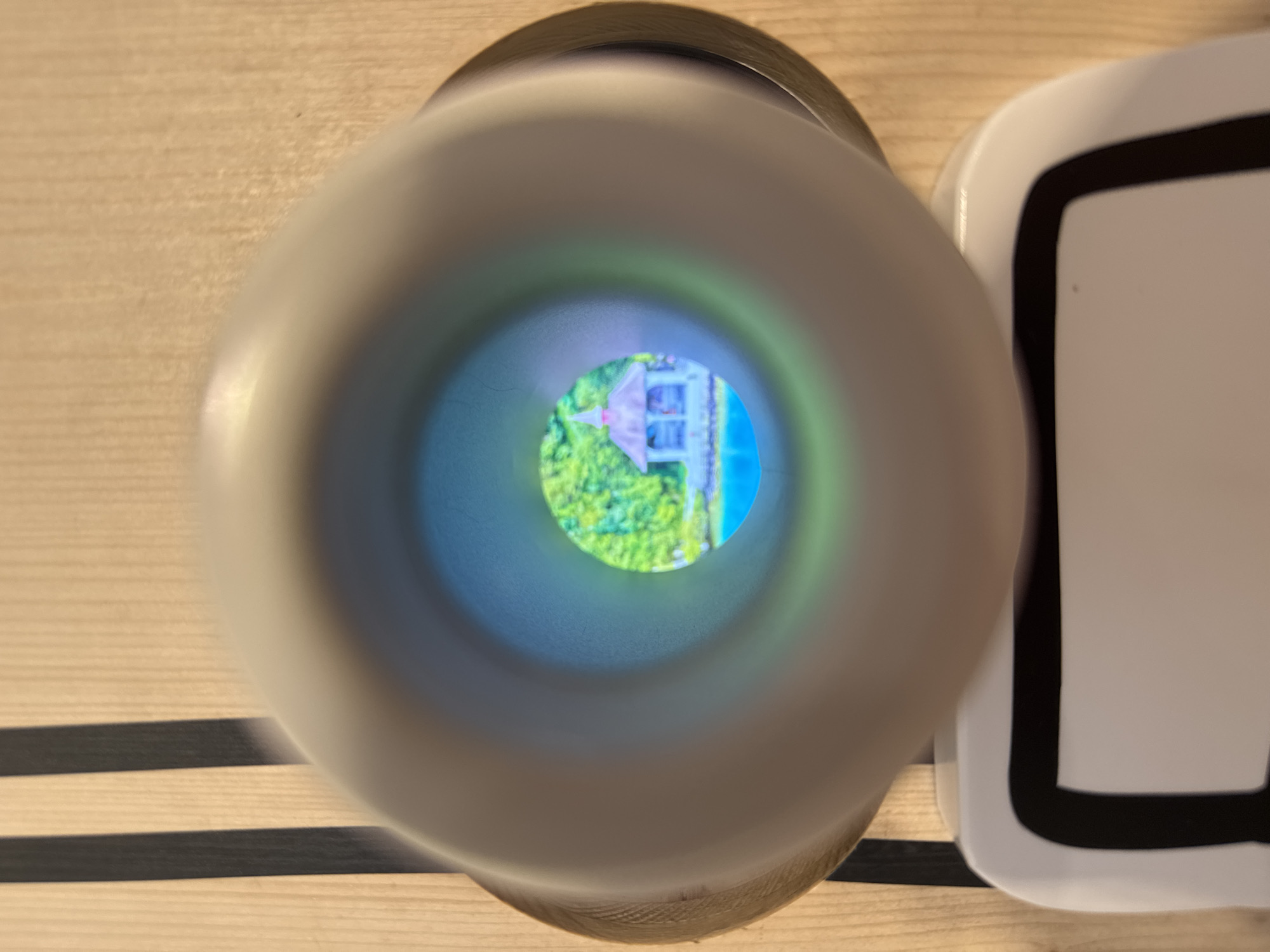 This image captures a close-up view of a peephole, through which a miniature scene of a gazebo beside a body of water is visible. The gazebo, with a pointed roof and visible pillars, appears to be located on a small island or peninsula. Lush green foliage surrounds the water, suggesting a natural, possibly park-like setting. The scene within the peephole is brightly lit and colorful. The outer elements of the image show the textured wooden surface of the object containing the peephole, with two vertical black stripes on the left side. A portion of a white object with black trim is visible at the bottom of the frame. This image is taken at the Uri Museum in Schattendorf, Switzerland, specifically in the "Museum im Ballade" exhibition, which features a diverse collection of objects, including those that offer unique perspectives or visual experiences.
