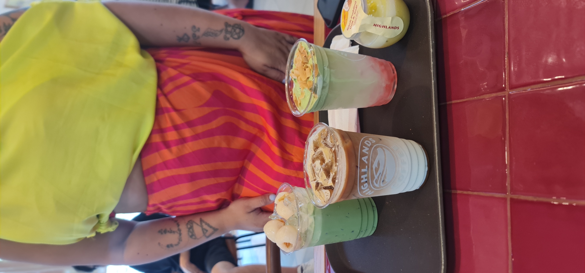 A person with arm tattoos wearing a yellow top and an orange and pink striped skirt is sitting at a table. On the table is a tray with three drinks and a small cup of Banh Pudding. The drinks include two iced beverages in clear plastic cups from Highlands Coffee, and one small cup with lychee. The tray is on a table next to a wall tiled with red glossy tiles. The image was likely taken in Đà Nẵng, Vietnam, as Highlands Coffee is a popular coffee chain in Vietnam.