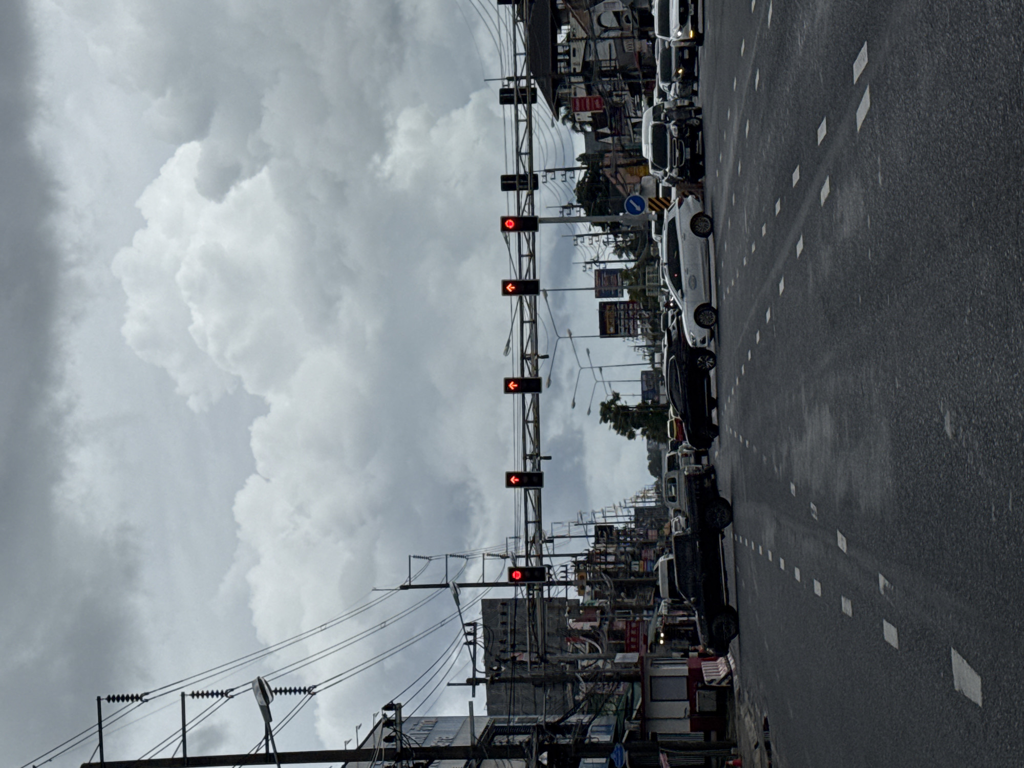 The image depicts a street scene in Ko Kaeo, Thailand, characterized by a busy intersection under a cloudy sky. Several vehicles, including a black pickup truck, a white SUV, a white van, and other cars, are visible on the asphalt road, some stopped at a traffic light. Traffic lights with red signals are illuminated, indicating a halt in traffic flow. The surroundings feature buildings lining the street, utility poles with numerous wires, and streetlights. The overall atmosphere is overcast, suggesting potentially inclement weather.