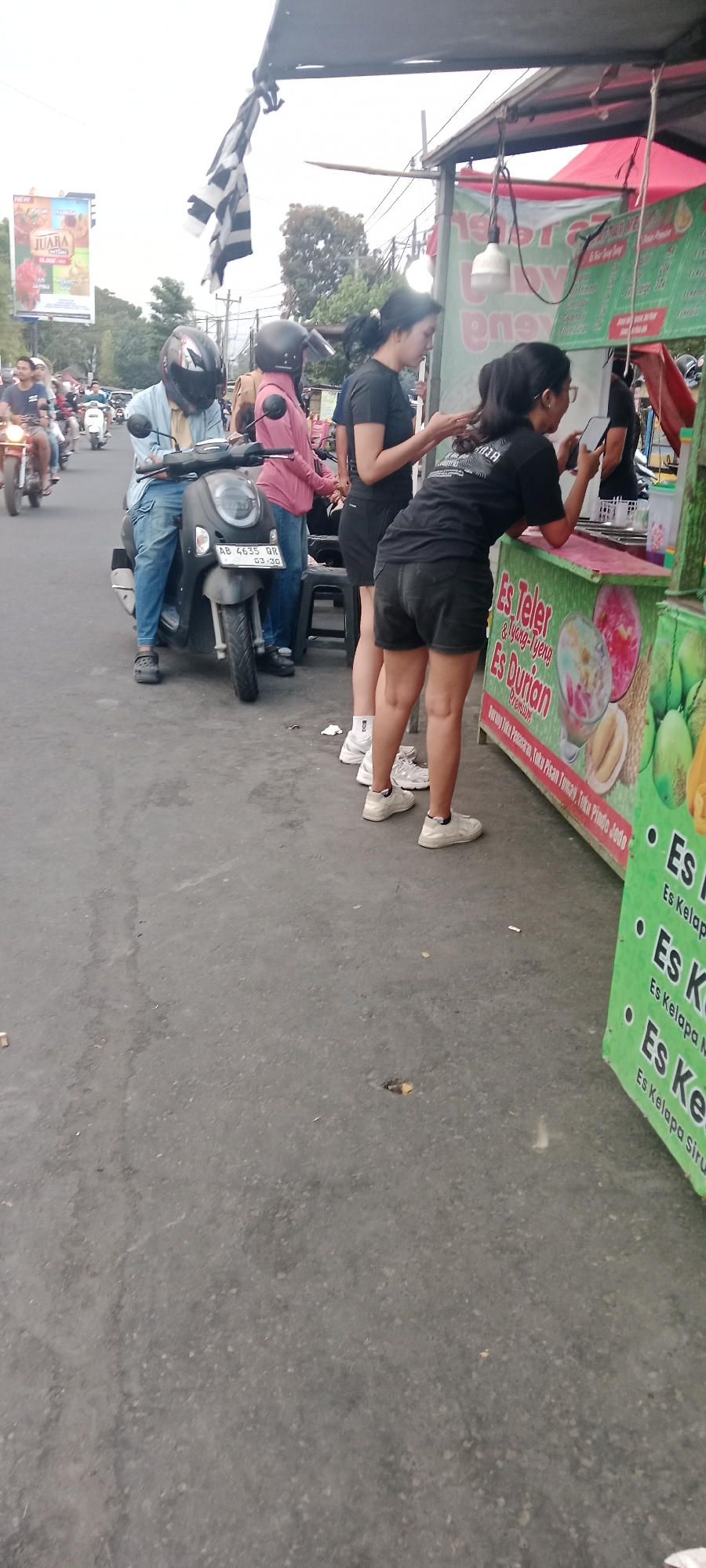 In Ngemplak, Indonesia, an outdoor scene depicts a street-level view with several people and a food stall. Two young women, one wearing a black t-shirt and dark shorts, and the other a black t-shirt and denim shorts, are standing at a colorful food stall that advertises "Es Teler & Yang-Uyeng Es Durian." The stall has a green and pink exterior with images of fruit and desserts. One woman is looking at her phone, while the other appears to be interacting with the stall. To the left, a person wearing a light blue shirt and jeans is seated on a dark-colored scooter, which has a license plate reading "AB 4635 QR." Another person in a pink long-sleeved shirt is standing next to the scooter. Several other individuals are visible on motorcycles further down the street. A large billboard with the word "JUARA" is in the background, along with trees and utility poles. The ground is paved asphalt, and there is some scattered debris. A black and white checkered flag is hanging from an overhead structure. The overall impression is of a busy street scene near a food vendor.