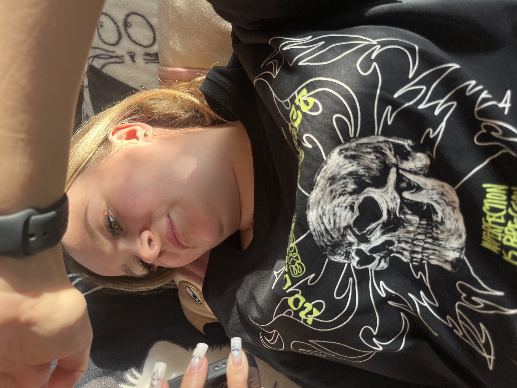 A selfie-style photograph presents a fair-skinned young woman with blonde hair. She is wearing a black sweatshirt featuring a white skull graphic and decorative flames. Her nails are long and painted white with a marble design. A black smartwatch is visible on her wrist. The background includes a blanket with a cat face design and a pillow. The image appears to be taken indoors. The location cannot be determined to be Podolsk, Russia, based on the visual information provided.