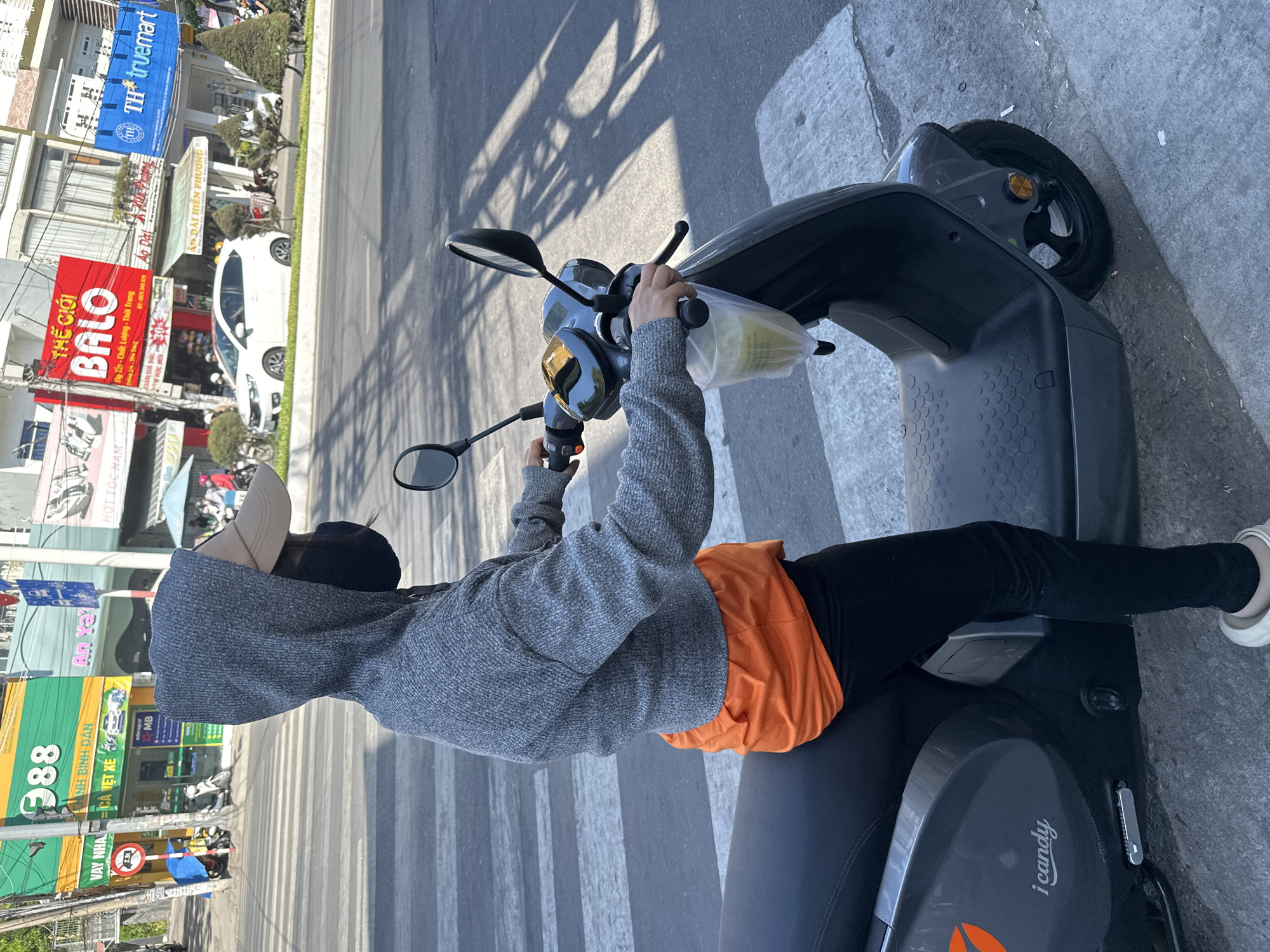 The image shows a person sitting on an "I Candy" brand electric scooter on a crosswalk. The person is wearing a gray hooded sweatshirt, an orange shirt underneath, black pants, a beige baseball cap, and a black face mask. They are holding the scooter's handlebars. The background includes street signs and storefronts in Bac Nha Trang Ward, Vietnam. Buildings and street signs, a white vehicle, and other parked motorbikes are in the background. The storefronts have signs indicating various businesses, including a backpack store ("THE GIOI BALO"), a bank (MB), and "TH truemart." A street sign shows "388" in green.