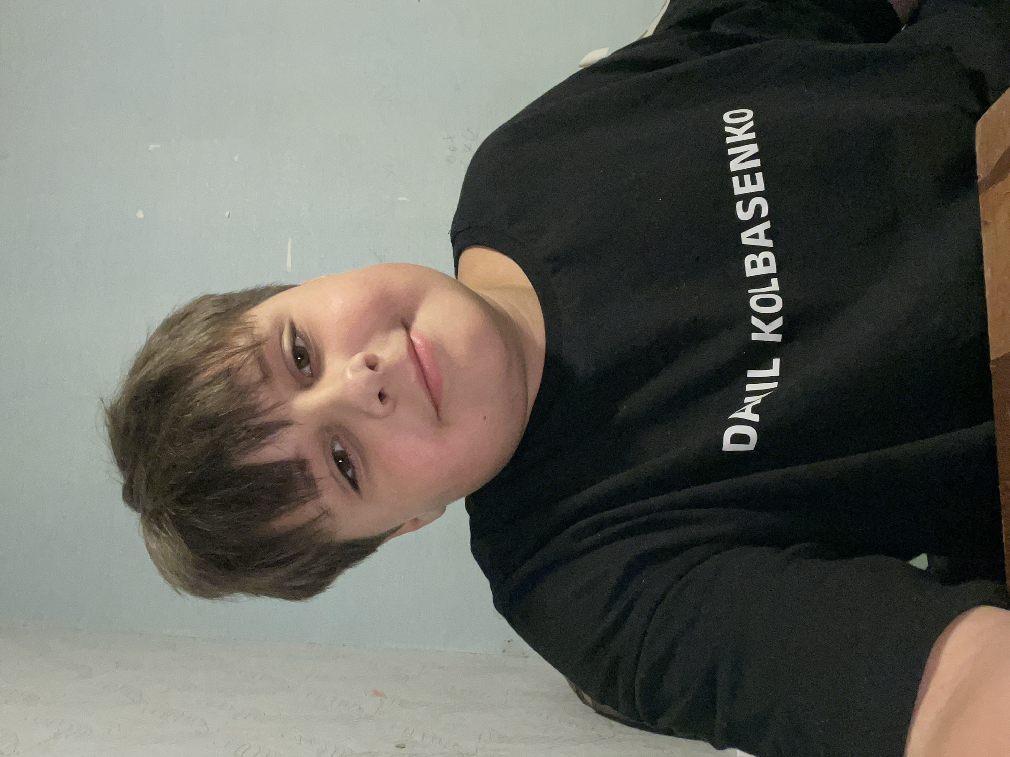 This is a portrait photograph of a young boy, seemingly indoors. The boy is positioned in the center of the frame, with his head tilted and his gaze directed towards the camera. He has brown hair and fair skin, with rosy cheeks. He appears to be wearing a black long-sleeved shirt that has white text printed vertically on the right side of the garment. The text reads "DAIL KOLBASENKO". The background consists of a light blue wall with some texture and a white, patterned surface below, possibly a floor or a table covered with a textured cloth. The lighting suggests it could be daytime, but no specific time is evident. The overall mood of the image is casual and personal, possibly a selfie taken by the boy. There are no clear indicators of the specific location beyond the general interior setting. The boy's expression is neutral to slightly smiling.