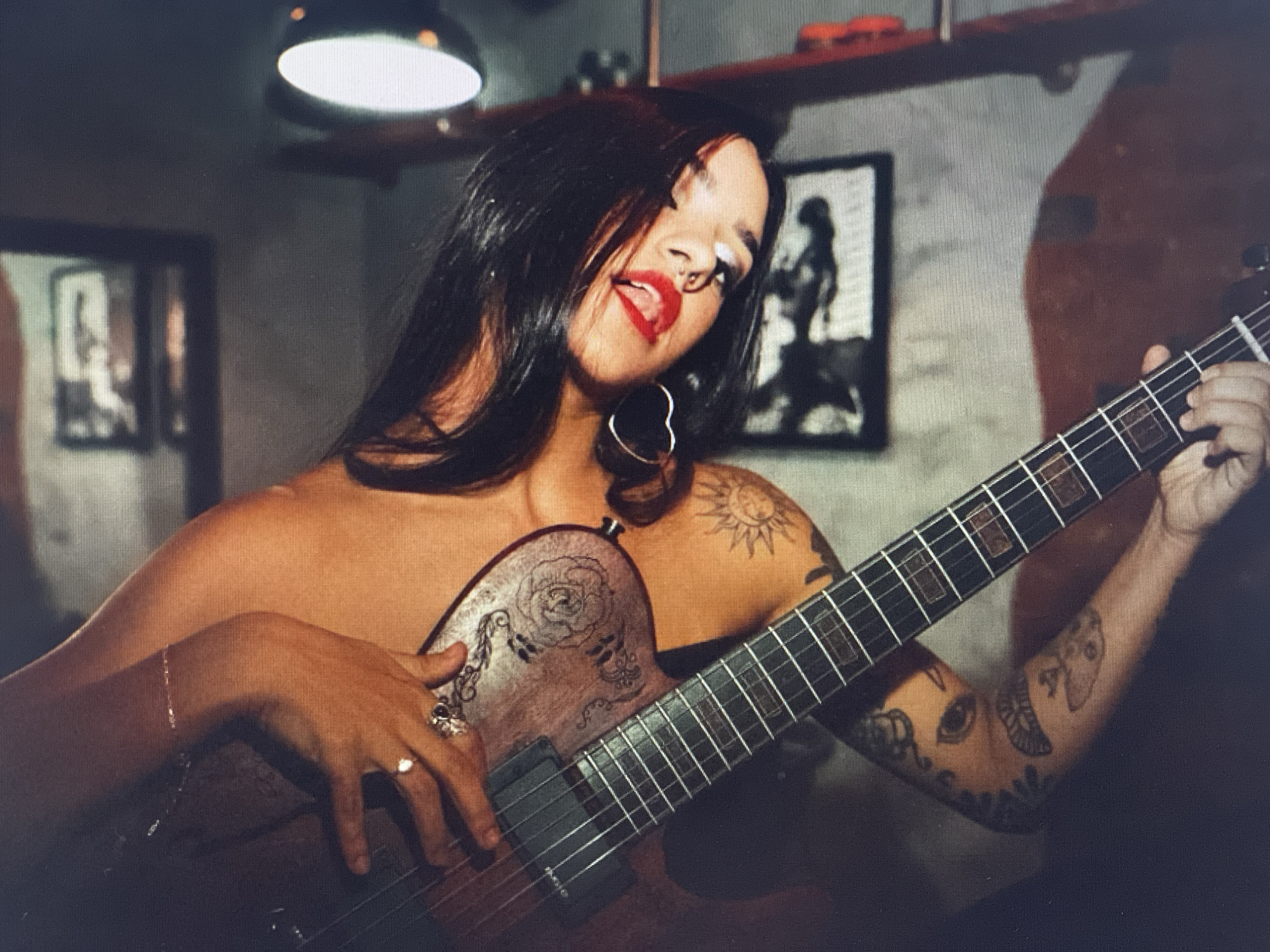 A woman with dark hair and red lipstick plays an electric guitar. She is looking into the camera with her tongue out. She has a nose ring and hoop earrings. Her left arm is resting on the guitar, and her right arm is extended, holding the neck of the guitar. She has tattoos on her arms and chest. The background is blurry, but appears to be a room with a framed picture on the wall and a lamp hanging from the ceiling. The lighting is dim.