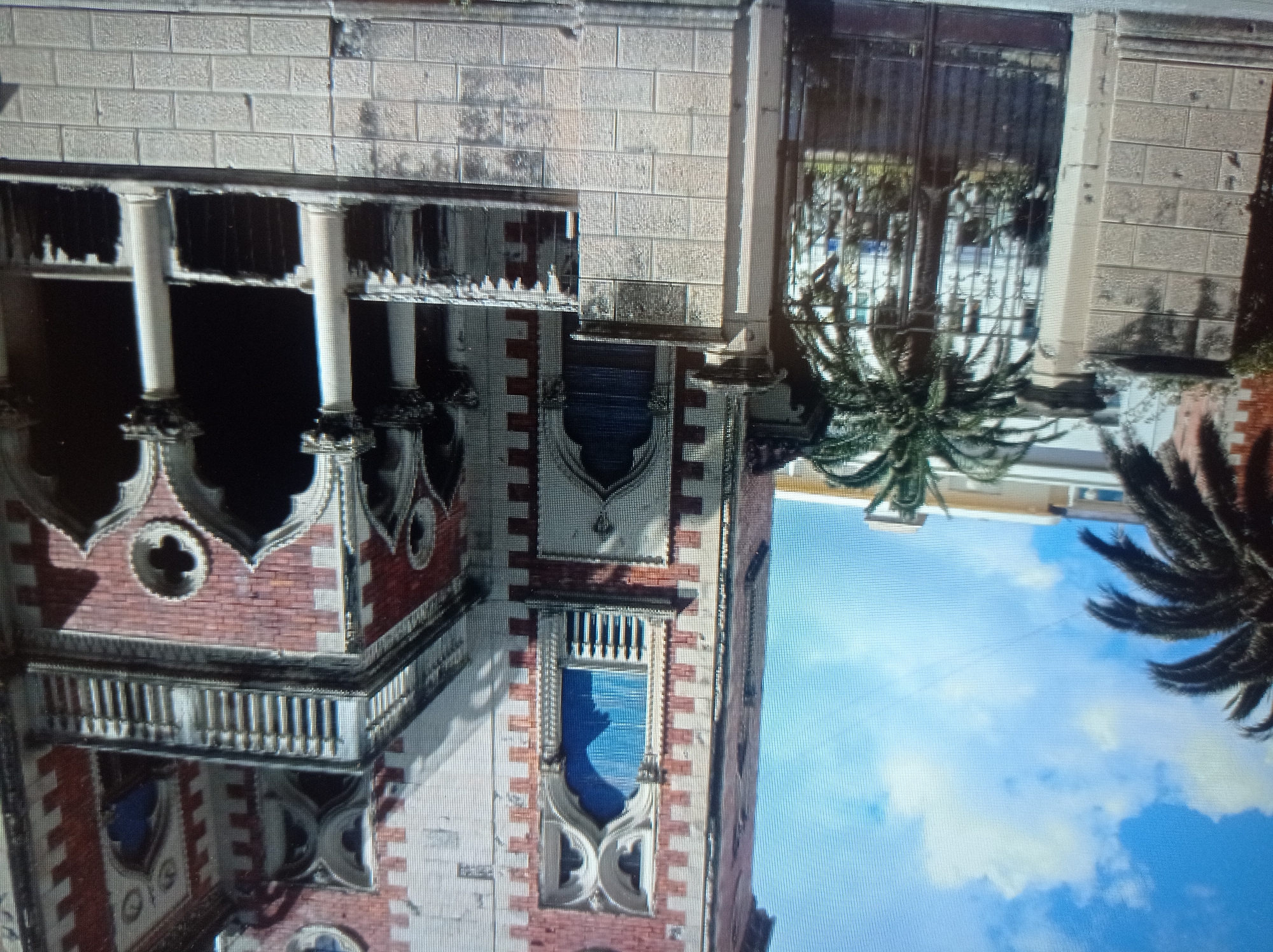 This image shows a red and white building with ornate architectural details, including arched windows and balconies with railings. Palm trees are visible in the foreground and to the left of the building. A stone wall and an iron gate are also present. The sky is blue with some clouds. The scene appears to be outdoors on a sunny day. The image does not contain any information that would identify it as being located in Mukachevo, Ukraine.