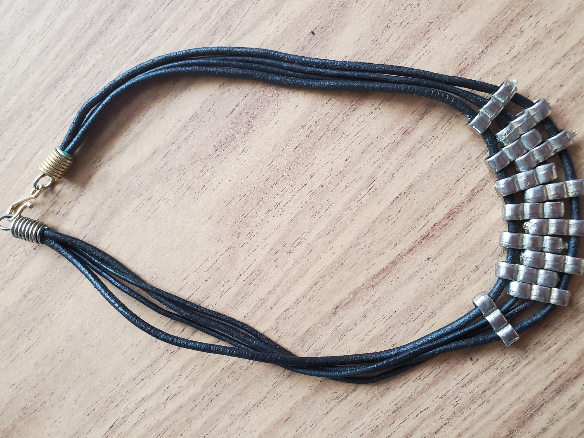 A close-up overhead shot shows a black leather multi-strand necklace with silver rectangular beads, resting on a light brown wooden surface. The necklace is laid out in a curved shape, with the clasp at the top right. The wooden surface has a visible grain pattern. There are no people or any indication of the location being in Tiraspol, Moldova.