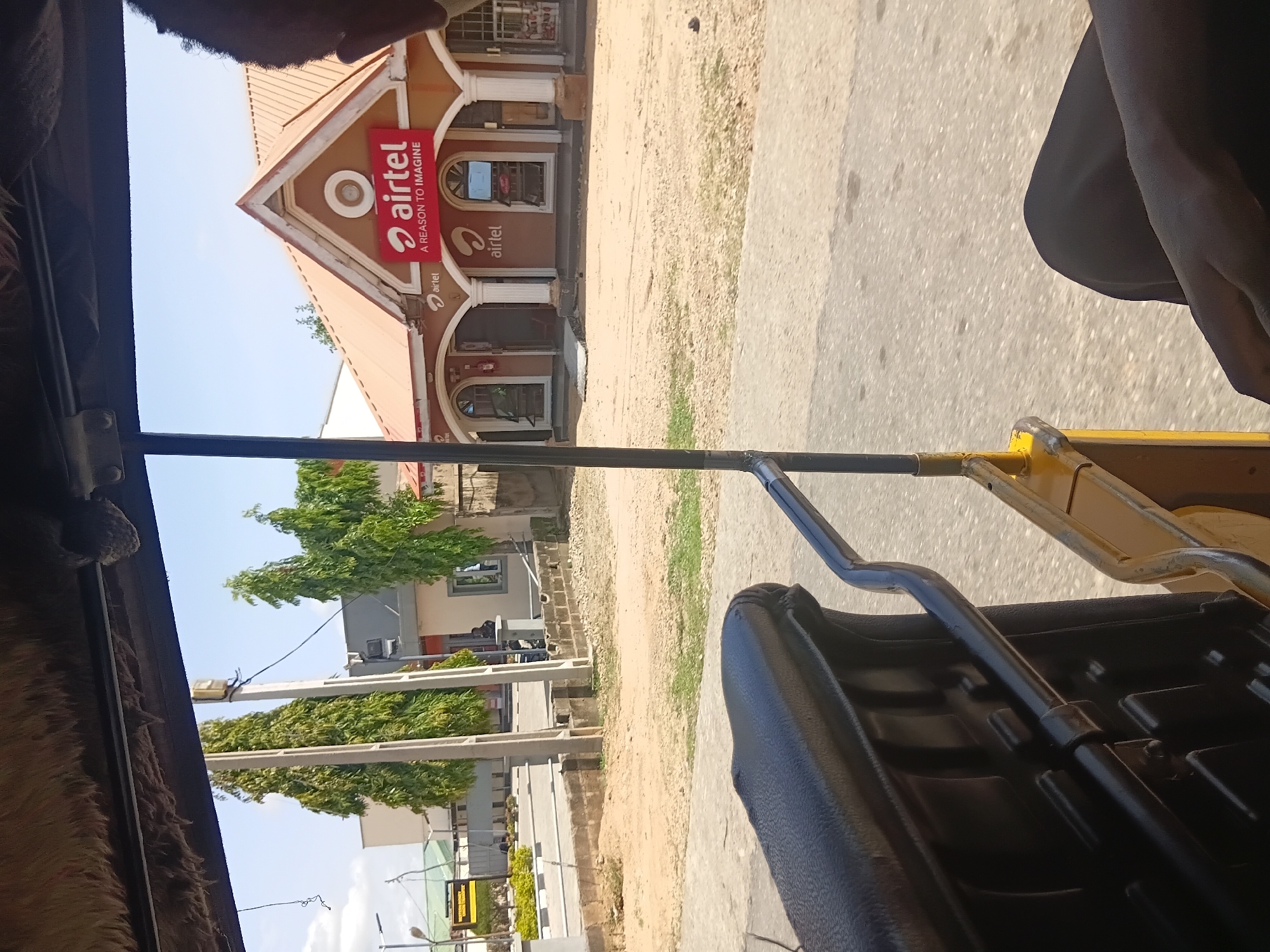 The image depicts a street scene in Jalingo, Nigeria, viewed from the interior of a motorized rickshaw. The rickshaw's weathered, fuzzy ceiling is visible at the top, along with its black seat and metal framework. In the background, an Airtel store, a telecommunications company, is prominently displayed with its red and white signage featuring the slogan "A REASON TO IMAGINE." The store has an orange roof and arched doorways, and several trees with lush green foliage line the street. Adjacent to the Airtel store, other buildings and structures are visible, including what appears to be a bank or office building with a green awning. The ground is a mix of sandy soil and pavement, with some patches of grass. The sky is clear and bright blue, indicating a sunny day.