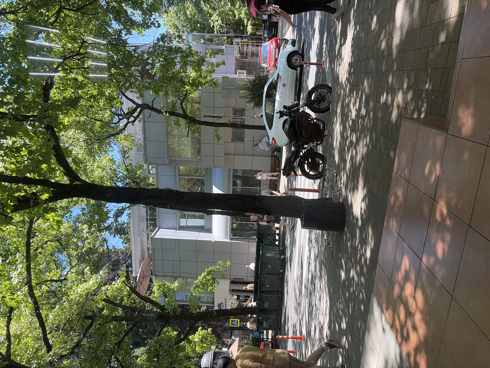 The image depicts a street scene in Sochi, Russia, on a sunny day. In the foreground, a tiled walkway is visible, with dappled sunlight casting shadows from overhead trees onto the tiles. To the right, a trash can with a wooden exterior stands on a gravel base. A black motorcycle is parked on the paved street.
Across the street, a modern, multi-story building with a grey and white facade dominates the background. Several large, green dumpster bins are lined up in front of the building. A white car with "МЕДСЛУЖБА" (medical service) written on its side and a red cross emblem is parked near the building. A red SUV and a black car are also visible further down the street.
Several people are present in the scene. A man in a tan shirt and shorts walks on the left side of the walkway. Other individuals are seen standing or walking near the building and the dumpsters, including a woman in a black top and skirt on the right side of the frame. A pedestrian crossing marked with white stripes is visible on the street. Orange traffic cones are placed along the edge of the walkway. Trees with lush green foliage line the street, casting shadows and contributing to the overall bright and summery atmosphere.
