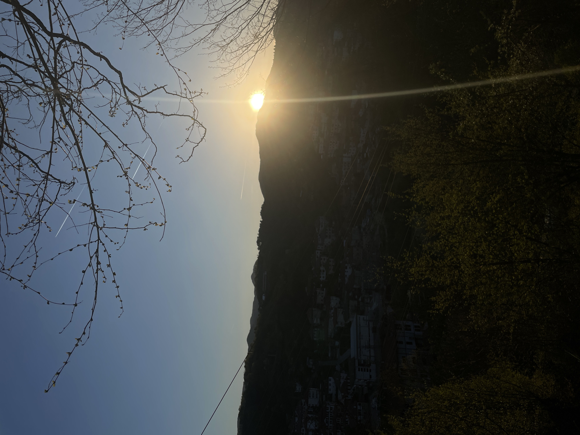 The image shows a landscape in Zlatograd, Bulgaria, featuring a mountain partially obscuring the sun. There are buildings situated on the mountain's slope. Bare tree branches with buds are visible in the foreground against a blue sky. Contrails from planes crisscross the sky. Power lines traverse the scene. Vegetation is also present on the right side of the image.