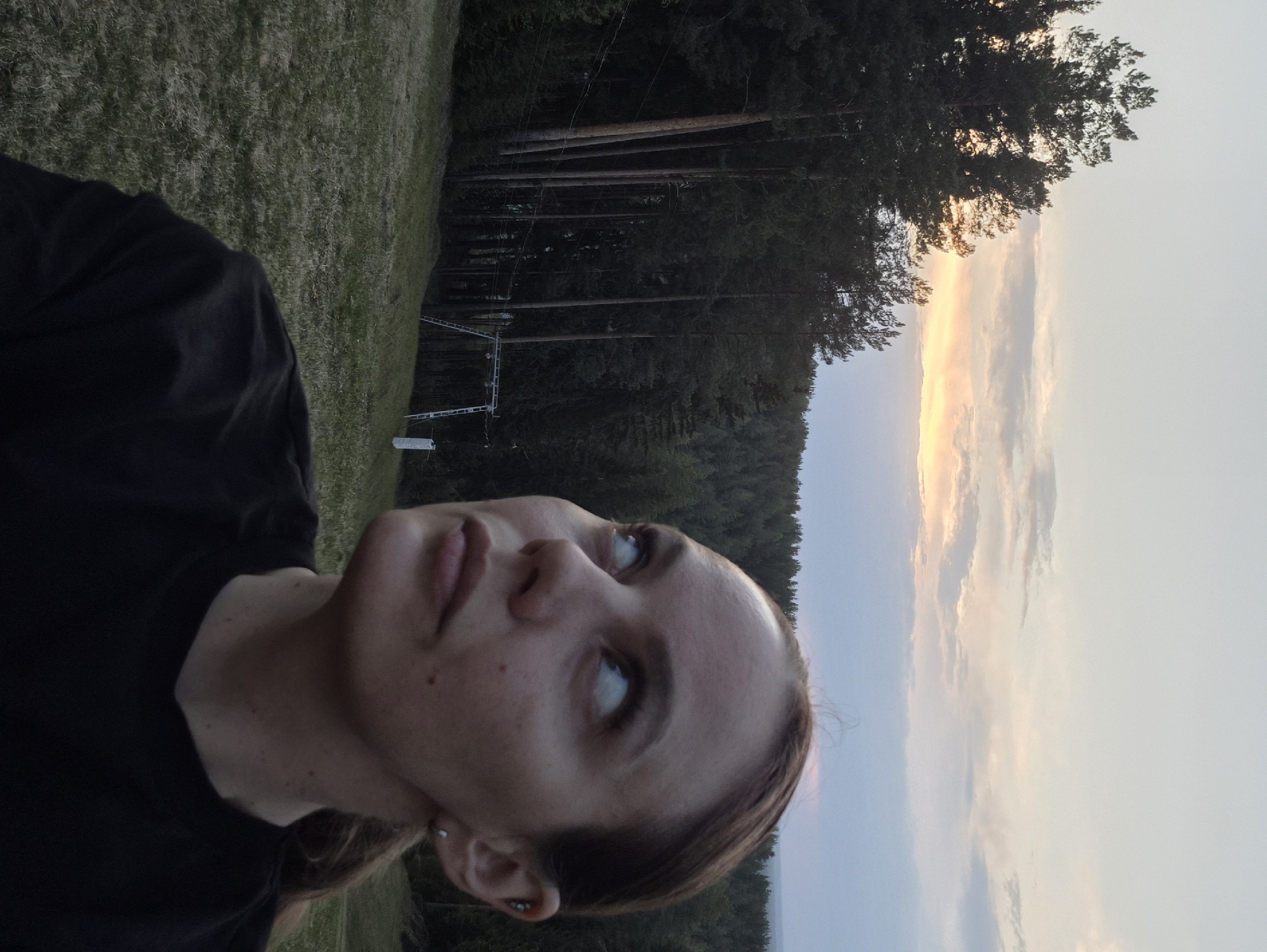 A woman is pictured in a close-up, looking upward and to the right of the frame. She has fair skin, with some freckles visible. She is wearing a black t-shirt. Her hair is pulled back. There appears to be an earring in her ear.
The background shows a grassy area, tall trees, and a sky with a prominent vertical band of light, suggesting either sunrise or sunset. There's a ski lift structure visible amidst the trees, pointing to the location possibly being a ski resort or similar recreational area within Прикамский, Russia.