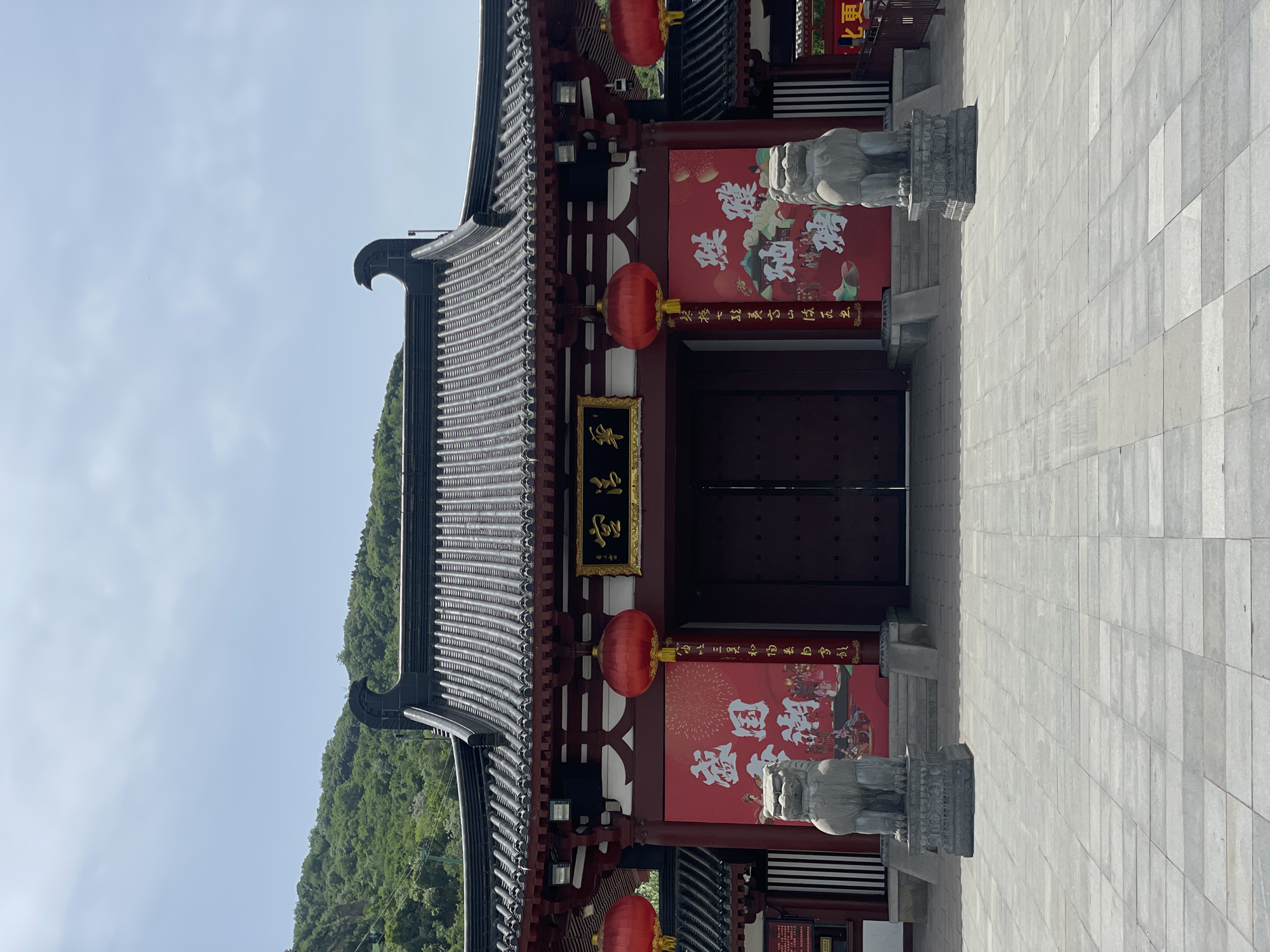 The image shows a traditional Chinese building in Lintong District, China, with a gray-tiled roof and red pillars. Two stone lion statues flank a set of large, dark wooden doors, adorned with red posters featuring white Chinese characters. Red lanterns hang on either side of the entrance. A sign with gold Chinese characters is placed above the door. A green, tree-covered hillside is visible in the background on the left, contrasting with the gray tiled courtyard in the foreground on the right, and the sky above.