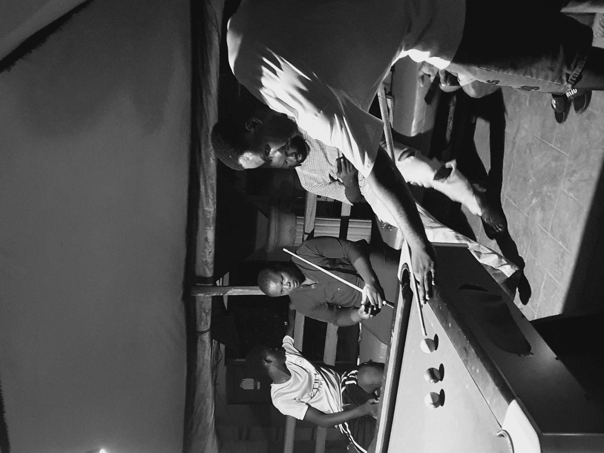 In Lokoja, Nigeria, four individuals are gathered around a pool table in an outdoor, dimly lit setting. The scene appears to be a casual gathering, possibly in a backyard or a recreational area.
In the foreground, on the right, a man is actively engaged in playing pool, holding a cue stick and aiming at the balls on the table. His body is angled towards the table, and his focus is on the game. To his left, another man, wearing a striped shirt, is seated and looking down, possibly at his phone or the game.
In the background, on the left, a young man in a t-shirt and shorts is leaning on the pool table, looking towards a woman seated on the other side. The woman, dressed in a dark top, is holding a pool cue stick in her hands, appearing to be either waiting for her turn or observing the game.
The pool table itself is the central object of focus, with several balls visible on its surface. The surrounding environment suggests a rustic or simple setup, with visible wooden beams and corrugated roofing overhead, typical of informal outdoor spaces in Nigeria. The lighting is subdued, creating a moody ambiance.