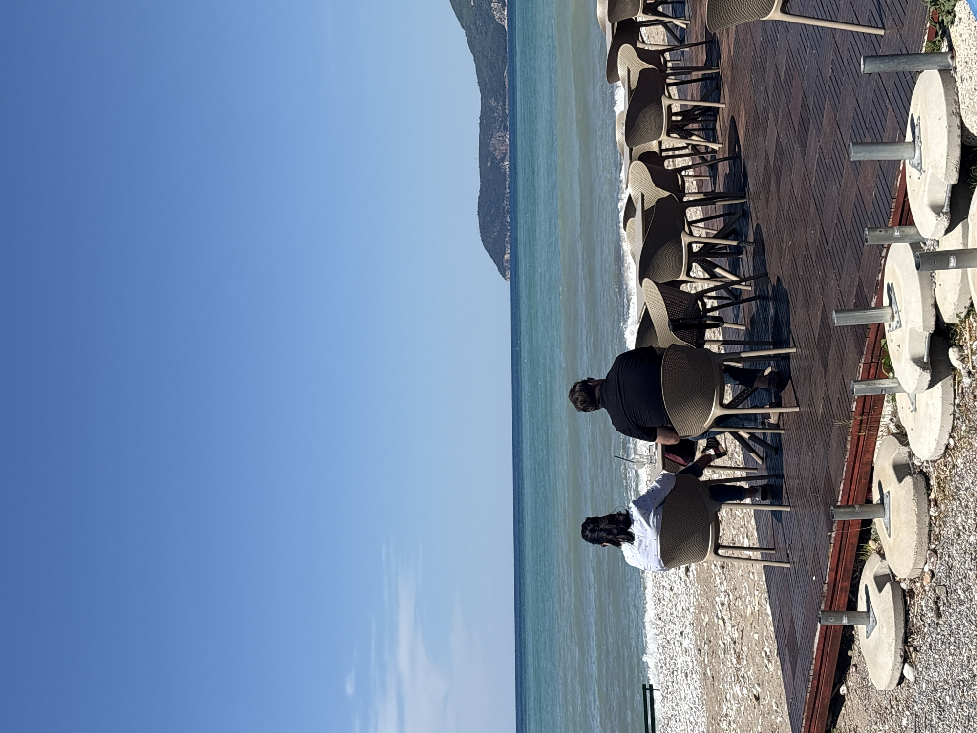 The image captures a vibrant, sunlit outdoor bar or restaurant patio situated directly beside the sea. Two individuals are seen from behind, seated on modern, dark-colored chairs at a table, facing the water. One person wears a dark shirt, and the other a white shirt with a subtle pattern. On their table, a glass with a straw and a small dark object (possibly a phone) are visible.
The patio features a dark wooden deck, populated by numerous empty chairs, indicating an outdoor dining area. To the right, off the main deck, there are several concrete bases with metal poles, likely for parasols, resting on a gravelly surface. Beyond the deck, a narrow pebble beach meets the calm, blue-green waters of the sea, with gentle waves breaking near the shore. A significant, dark mountain range stretches across the horizon in the distance, rising directly from the sea.
The scene is bathed in bright daylight under a clear blue sky, suggesting a sunny day, likely mid-day or early afternoon, given the distinct shadows cast by the chairs and people on the wooden deck. The overall impression is one of relaxation and enjoyment in a picturesque coastal setting. No discernible text is visible in the image. The setting is consistent with a bar in a coastal town like Bar, Montenegro.