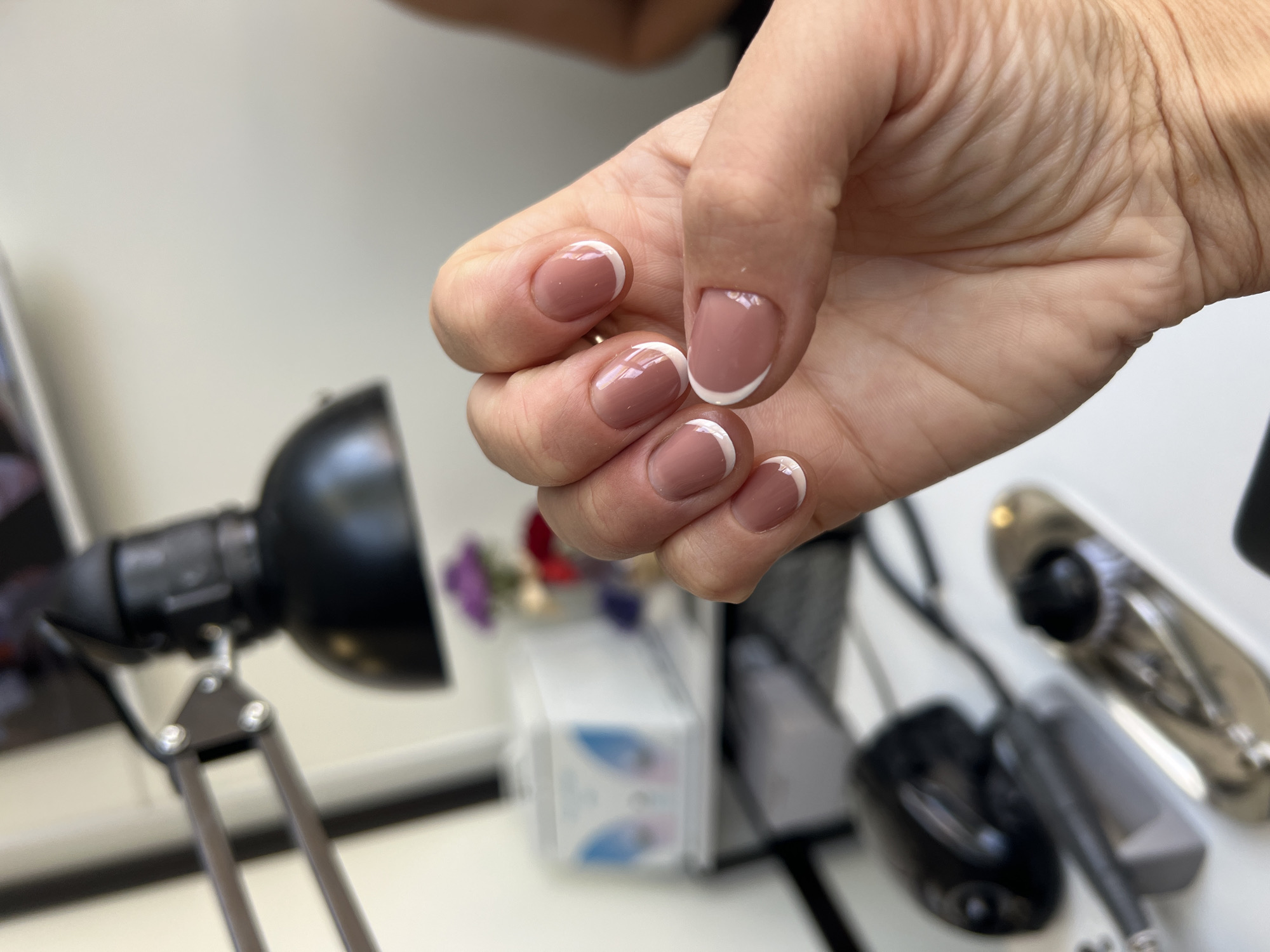 A close-up shot shows a person's hand with a fresh manicure featuring a nude base coat and white tips, commonly known as a French manicure. The nails are neatly shaped and polished, with a glossy finish. The hand is positioned in front of a blurred background that suggests a nail salon or a workspace. Visible in the background are various tools and equipment associated with nail care, including a black desk lamp with an adjustable arm, what appears to be a nail drill, and a white box with blue detailing, possibly containing nail supplies. The overall scene is well-lit, highlighting the detail of the manicure. The context of Zolotonosha, Ukraine, is not directly evident from the visual information in the image itself, but the setting appears to be an indoor establishment.