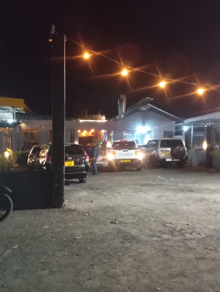 The image shows a parking lot at night in Arusha, Tanzania. There are five vehicles parked in the lot: a black SUV, a silver SUV, a white pickup truck, and two other cars. The vehicles are parked facing towards the camera. The white pickup truck has the word "ISUZU" written on the back. The black SUV has a license plate that is visible in the image. There are two people walking in the background, both wearing dark clothing, near the white pickup truck. There is a sign above the white pickup truck with the word "Shambe" on it. There are several string lights strung across the top of the parking lot. There is a street light illuminating the area in the background. The parking lot is made of gravel. There is vegetation in the background of the image to the right. The image is captured from a low angle looking up towards the parking lot. The location of the parking lot is a business district in Arusha, Tanzania.