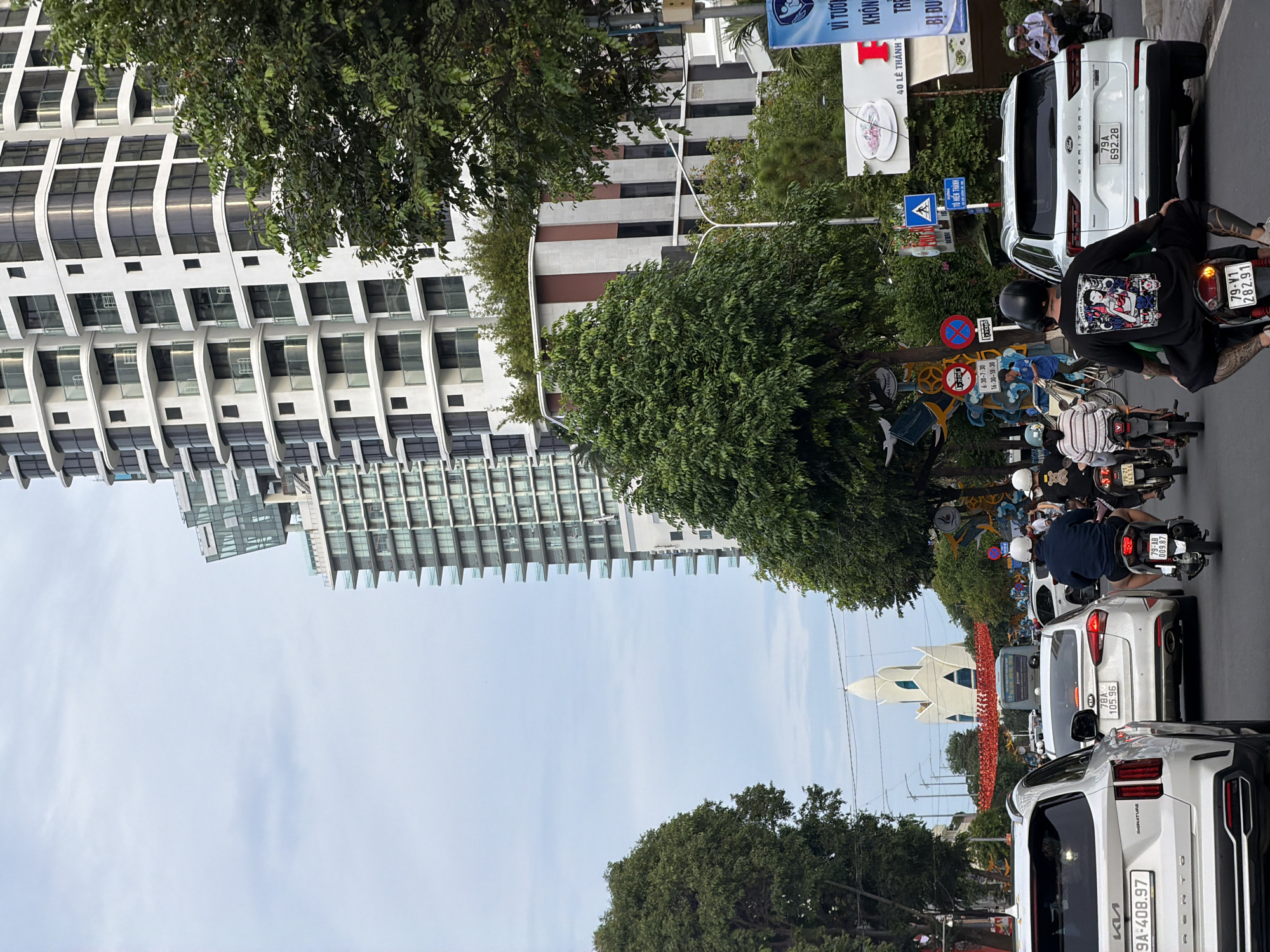 This image captures a vibrant street scene in Nha Trang, Vietnam, during daytime under a partly cloudy sky. The street is bustling with traffic, primarily motorbikes and several cars, suggesting a busy period.
In the foreground, a person on a motorbike, wearing a black helmet and a black hoodie with a colorful graphic print, is visible. This rider also has prominent tattoos on their legs. Other motorbikes carry multiple passengers, most wearing helmets. White SUVs are interspersed in the traffic, including a Kia Sorento (license plate 9A-408.97) and another SUV with license plate 79A 692.28. Motorbike license plates 79-V1 282.91 and 79-AB 009.87 are also visible.
Along the left side of the street stands a tall, modern high-rise building with a distinctive white facade featuring both curved and rectangular windows. Further in the distance, another multi-story building with a more grid-like window pattern is seen. Lush green trees line the street and appear in medians, providing a contrast to the urban architecture.
Various street signs are present, including a blue directional sign pointing to "TÔ HIẾN THÀNH," a large white billboard displaying "40 LÊ THÁNH" and a graphic of a white cup, and several traffic restriction signs indicating specific times: "6:30-7:30" and "16:30-18:30." A "no parking" sign is also visible. Other partial text includes "VÌ TƯƠI," "KHÔNG TRE," and "BI DU."
In the middle ground, beyond the immediate traffic, a unique, angular white monument or building with multiple peaks stands out against the sky. In front of it, a large, red, decorative art installation or display adds a splash of color to the scene. The overall impression is that of a dynamic and active urban environment.
