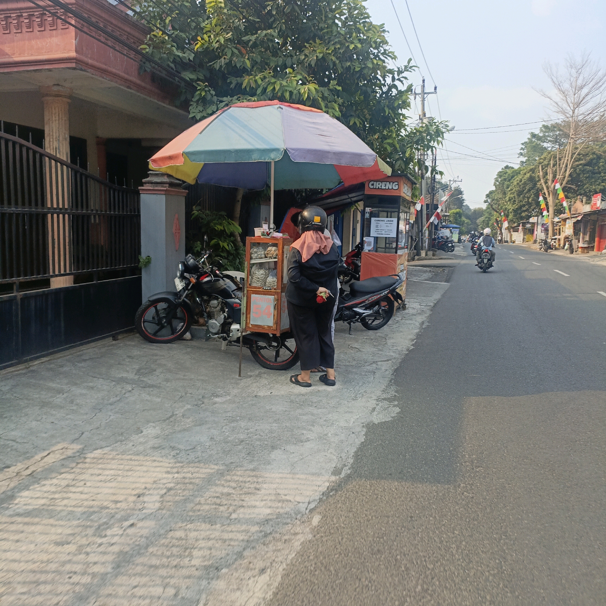 A woman wearing a hijab and black clothing stands behind a food cart. The cart has a colorful umbrella and displays food items in clear bags. A motorcycle is parked next to the cart, and another motorcycle is parked behind it. The scene appears to be on a street in Salatiga, Indonesia, with a building on the left and a road with passing motorcycles on the right. The street is lined with shops and flags. The ground is paved, and there are shadows cast by the objects and trees. The overall lighting suggests it is daytime.
