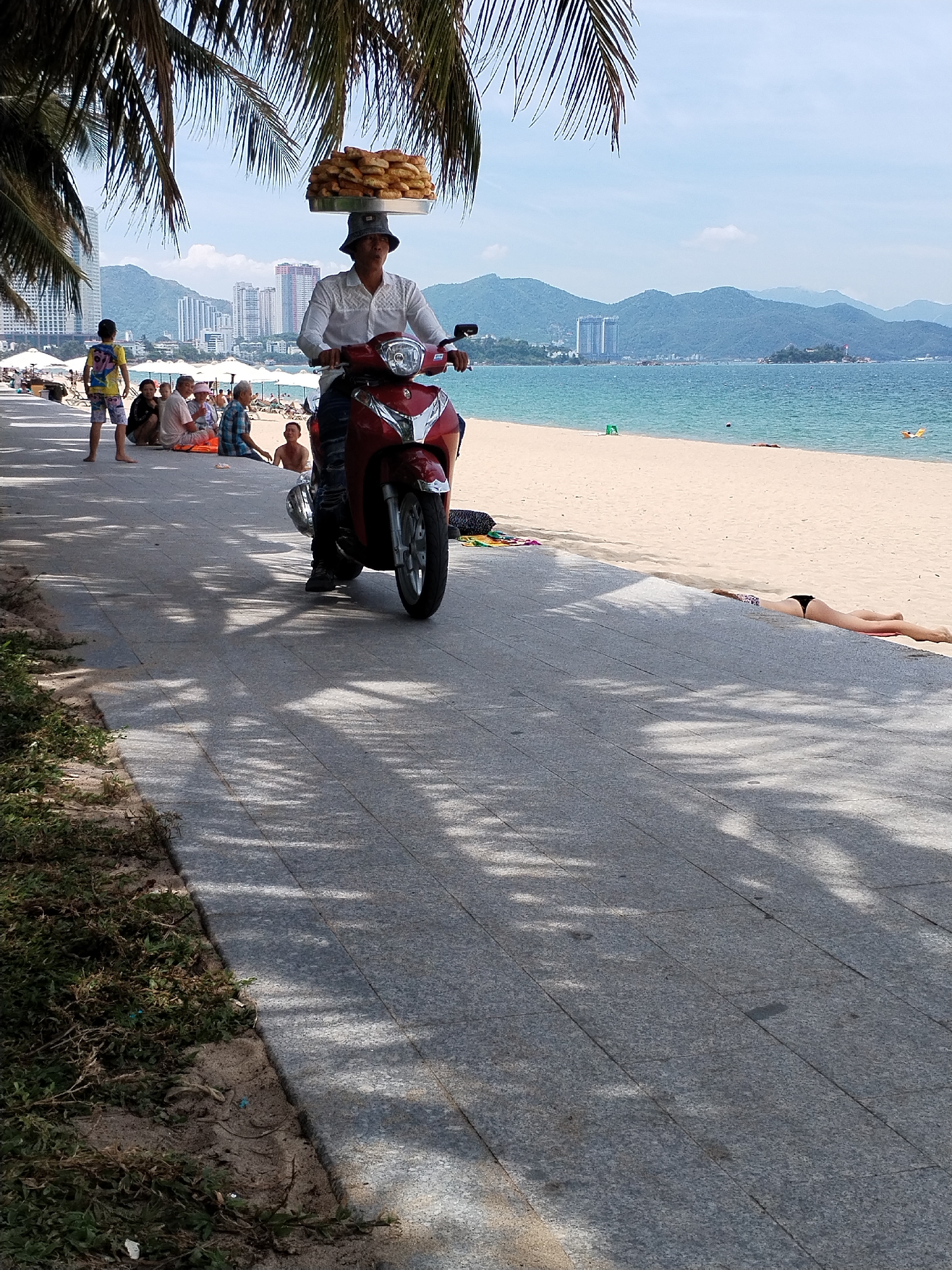 A vendor on a red scooter carries a large tray of food on his head while riding along a paved walkway next to a beach. The vendor is wearing a white shirt and a hat, and the food appears to be bread or pastries. Several people are sitting on the sand and a few are in the water. The background shows a city skyline with mountains. The image was taken in Nha Trang Ward, Vietnam.