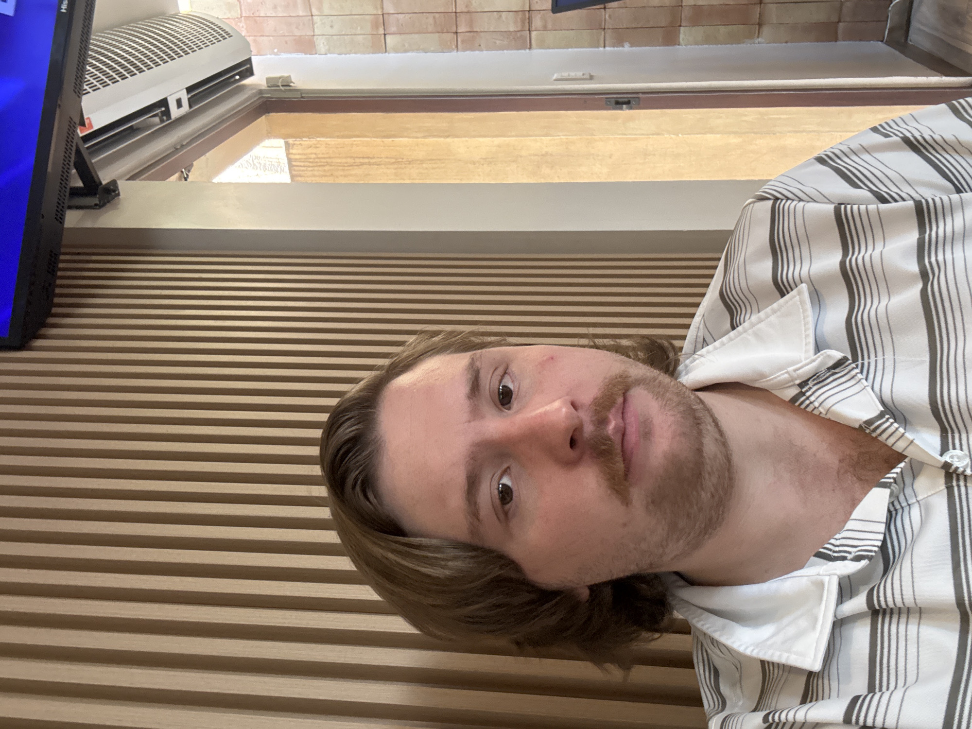 A sideways head-and-shoulders shot of a man with light brown hair and a mustache wearing a striped shirt. He is positioned in front of a wall with horizontal wooden slats. In the background, a window or skylight allows daylight to enter, above which is a white air conditioning unit. The location is presumed to be in Cuautitlán Izcalli, Mexico.