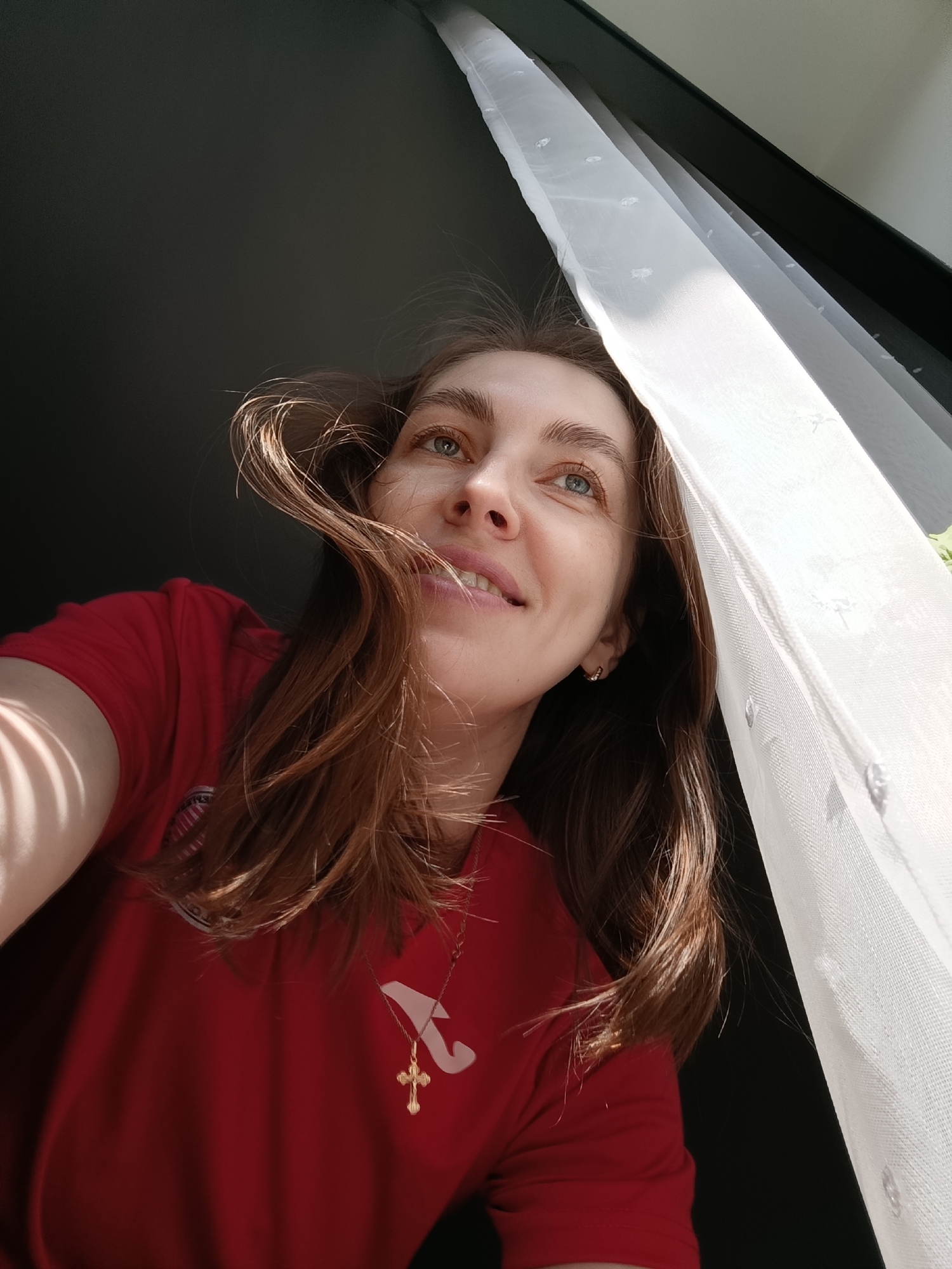 A low-angle, eye-level shot features a young adult woman looking upwards and slightly to the right with a soft, content smile. Her long, medium-brown hair is gently windblown, framing her face. Bright, natural light illuminates her from the right, highlighting her blue eyes and fair complexion. She wears a red t-shirt, which has a white stylized 'A' logo and the word 'JOMA' visible on the left side of her chest. A golden chain with a golden cross pendant adorns her neck, and a small hoop earring is visible in her right ear.
To her right, a white, sheer curtain hangs, appearing translucent as bright sunlight filters through it, revealing a subtle patterned texture. The curtain and an unseen window frame are the primary source of light, casting soft shadows on the woman's left arm. The background behind her is dark and indistinct, suggesting an indoor setting with strong, directional daylight. The time of day appears to be daytime, with ample sunlight. The woman's expression conveys happiness and contentment.
