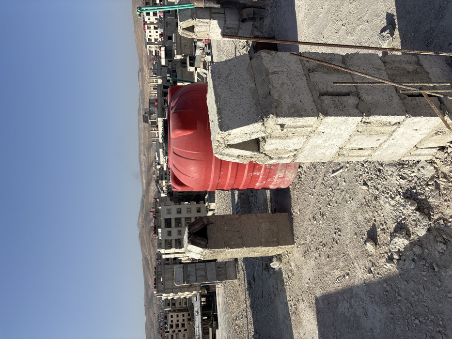 The image depicts an elevated view of a cityscape under a clear blue sky in Mu'adammiyat ash-Sham, Syria. In the foreground, a dusty, gravel-covered rooftop features several unrendered concrete block structures, one of which has a distinct triangular top. A large, red, cylindrical plastic water tank stands prominently in the mid-ground. Near the concrete structures, several rusty metal rebar rods protrude vertically from the ground. The urban environment extends into the background, showcasing numerous multi-story residential buildings, many with visible water tanks on their rooftops and some appearing unfinished or under construction. Distant, arid hills frame the horizon, with a faint plume of smoke or haze visible on the left side. No individuals are present in the scene. The lighting suggests bright daylight, casting strong shadows from the foreground objects.