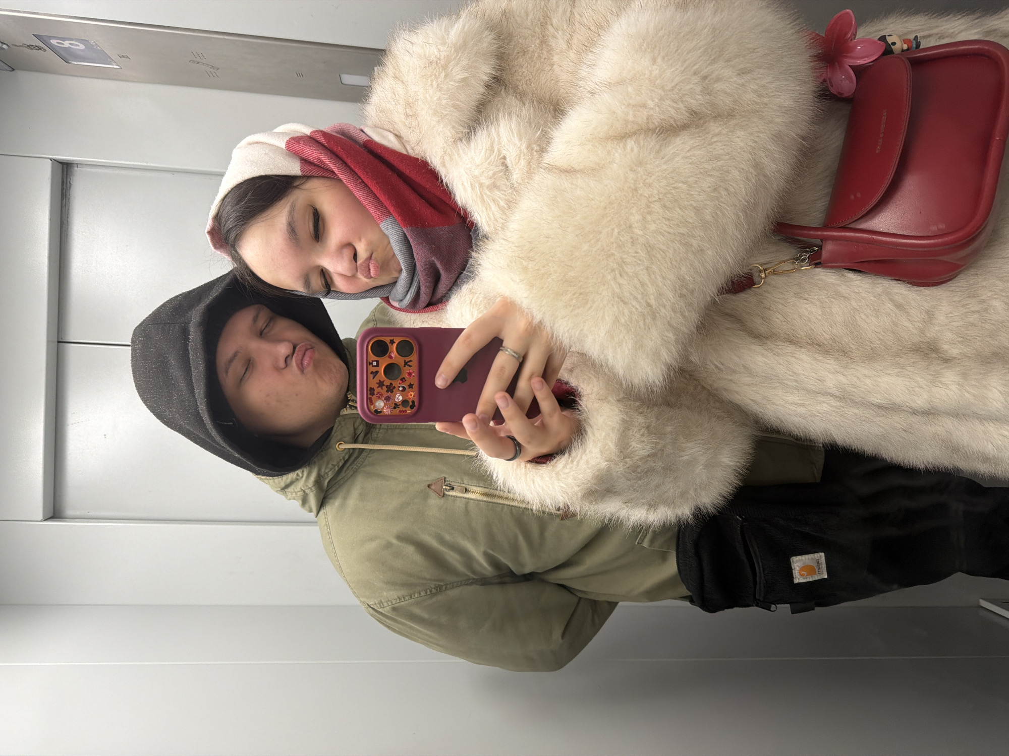 Two individuals are captured in a selfie within an elevator, likely in Perm, Russia, based on metadata. The person on the right wears a white fur coat with a red purse adorned with a flower and cartoon character. A red and white scarf covers their head. The person on the left wears a green hooded jacket with a dark hood pulled up. Both are making a kissing face toward the camera. The person on the right holds a maroon-colored iPhone with a decorated case, indicating they are taking the photo. A black Carhartt bag is visible on the person wearing the hooded jacket. The interior of the elevator is visible, featuring gray metal panels and a floor indicator.