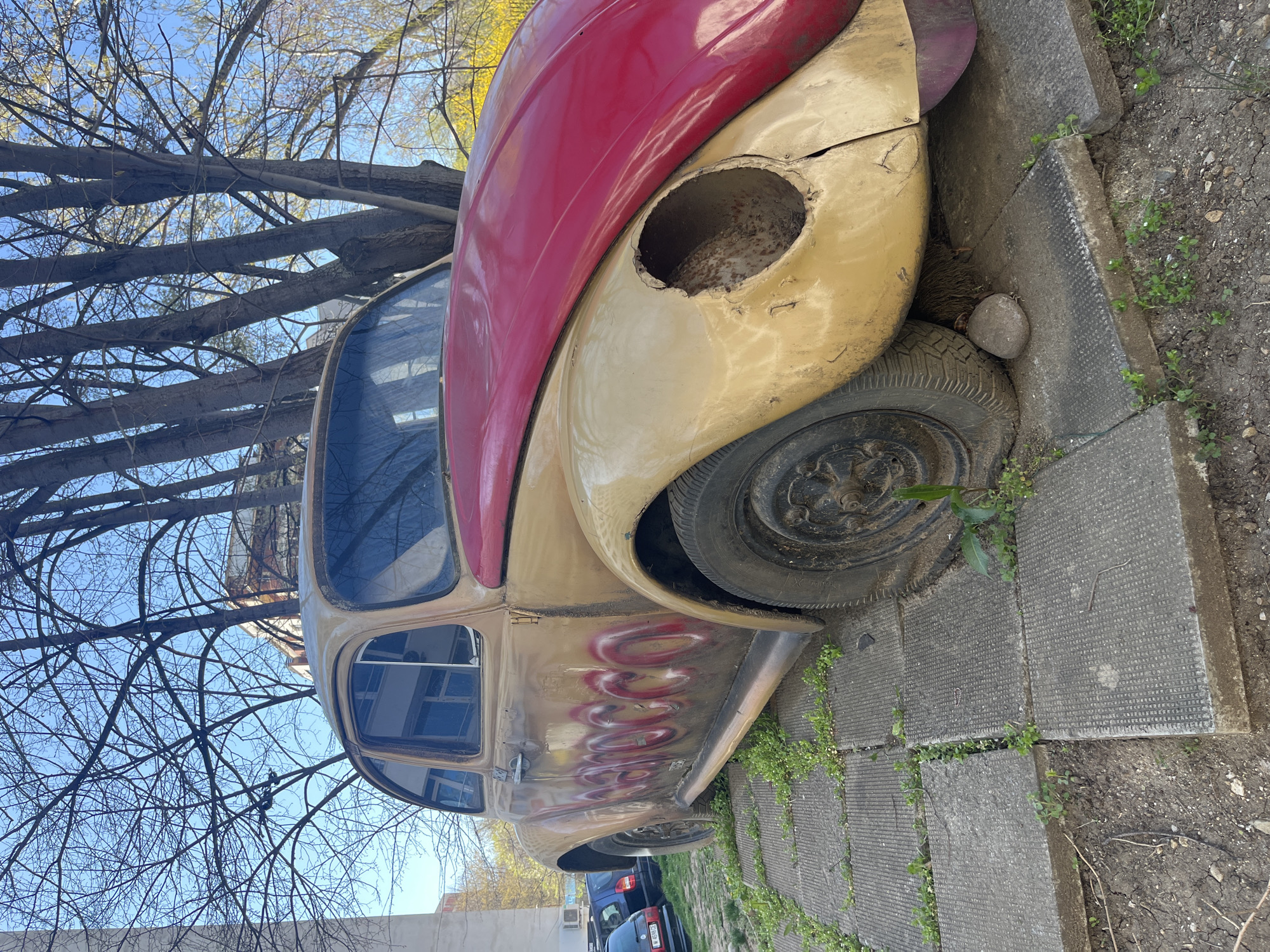 An aging, stationary, two-toned (red and faded yellow) vehicle, possibly a car, rests partially on a paved concrete surface, bordered by dirt and sparse green vegetation. A round opening is visible in the front fender where the headlight once was. The car is positioned near trees with bare branches against a blue sky. In the background, other cars are visible. The location is identifiable as the city of Sofia, Bulgaria. No people are visible. The car appears to be abandoned or in disrepair.
