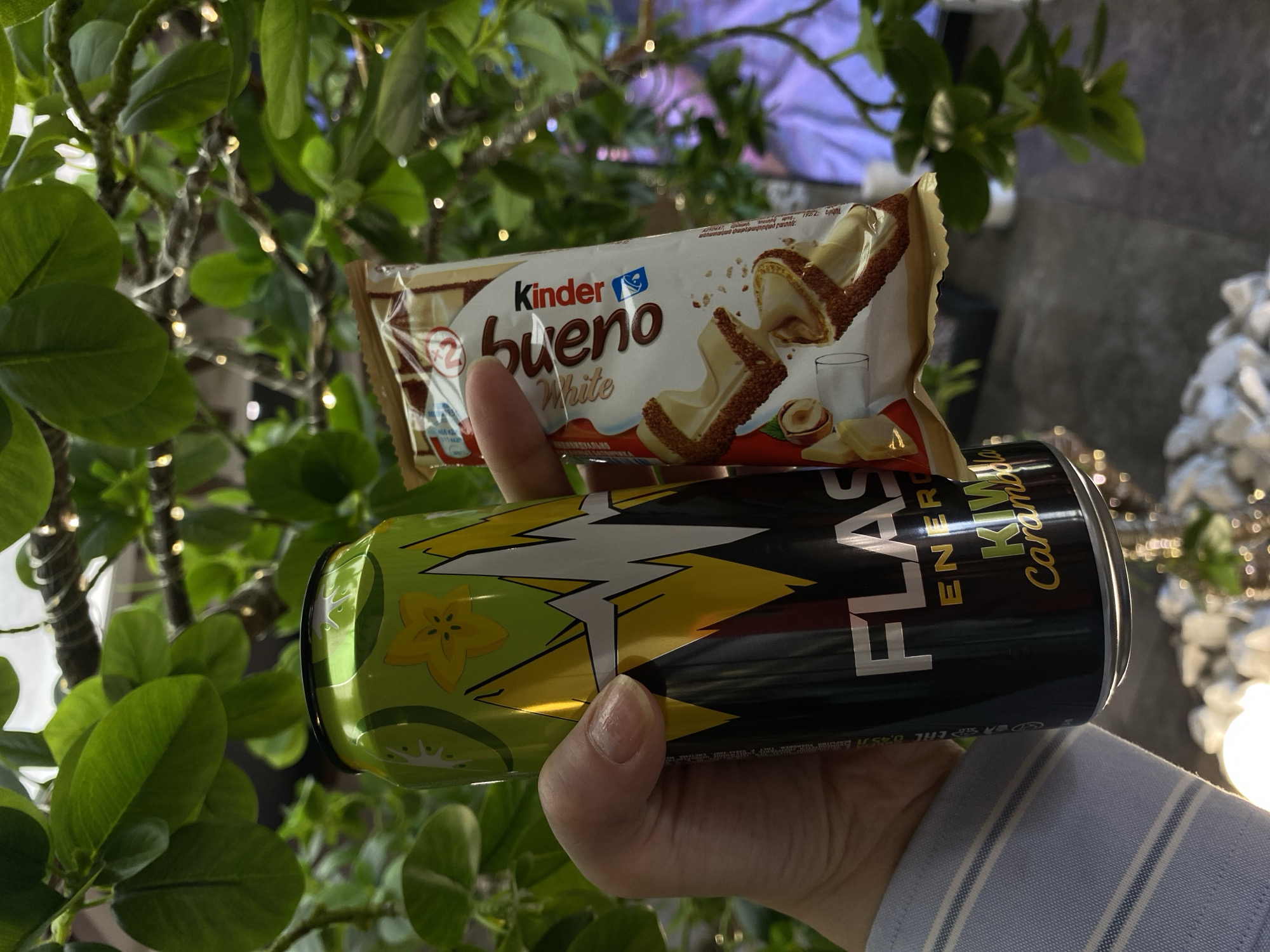 A hand holds a can of Flash Up Energy Kiwi Caramel flavored drink and a Kinder Bueno White chocolate bar. The background includes a leafy plant with small white lights. The location is unknown, and there's no information to indicate Votkinsk, Russia.