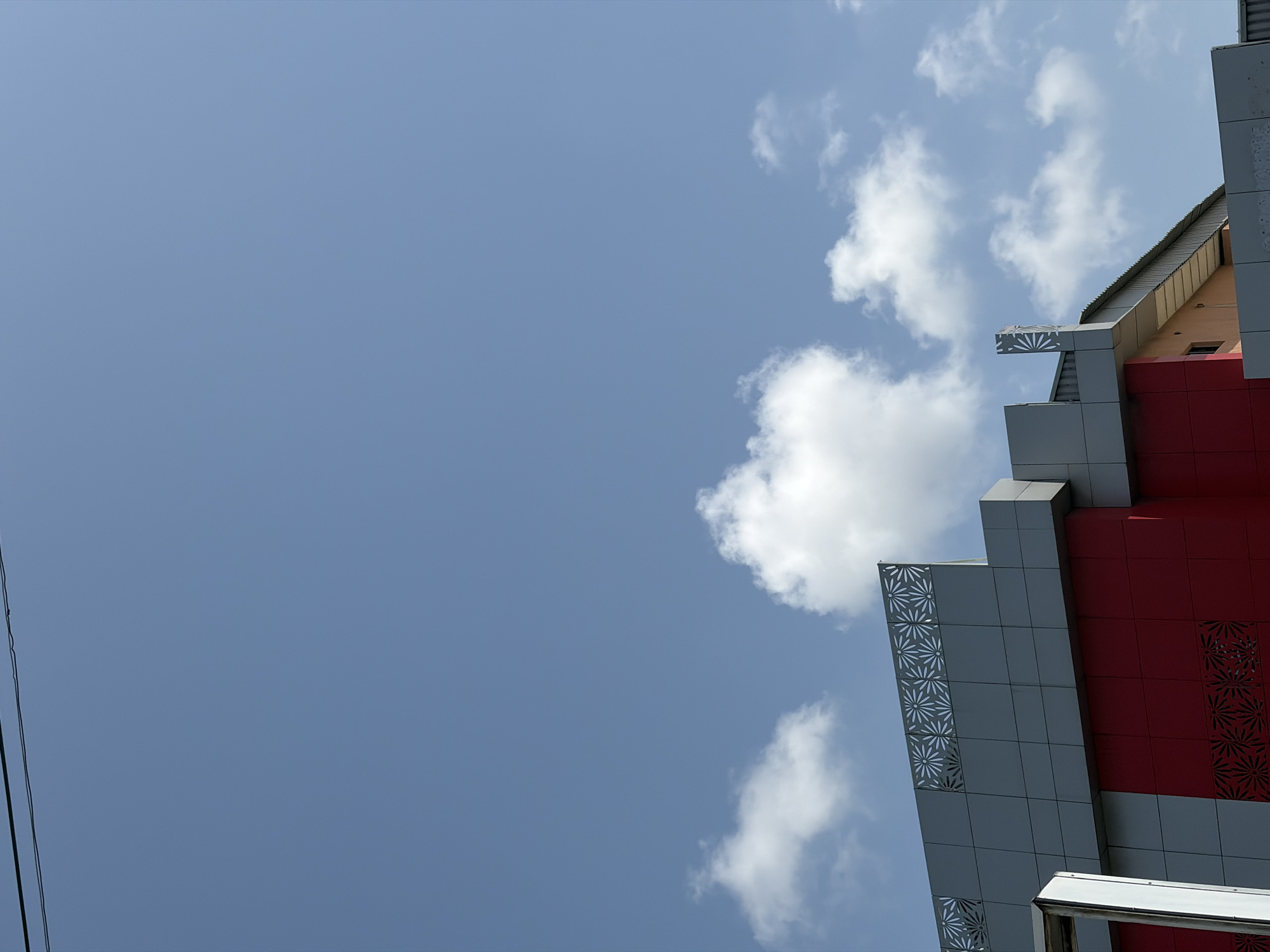 A low-angle, upward-facing shot captures a modern building against a clear blue sky with scattered white clouds. The building, located in Igboefon, Nigeria, features a distinctive facade primarily composed of gray and red panels. Several gray panels are adorned with intricate cutout patterns resembling stylized flowers or starbursts, adding an ornate touch to the contemporary design. The red panels are a prominent feature, contrasting with the gray. Towards the top, a roof structure and a small, light-colored section with a window are visible. The bright blue sky and fluffy white cumulus clouds suggest a clear, sunny daytime. On the far left, thin black power lines stretch vertically into the frame. There are no people visible, and no specific activity is taking place. No text is visible on the building or in the sky.