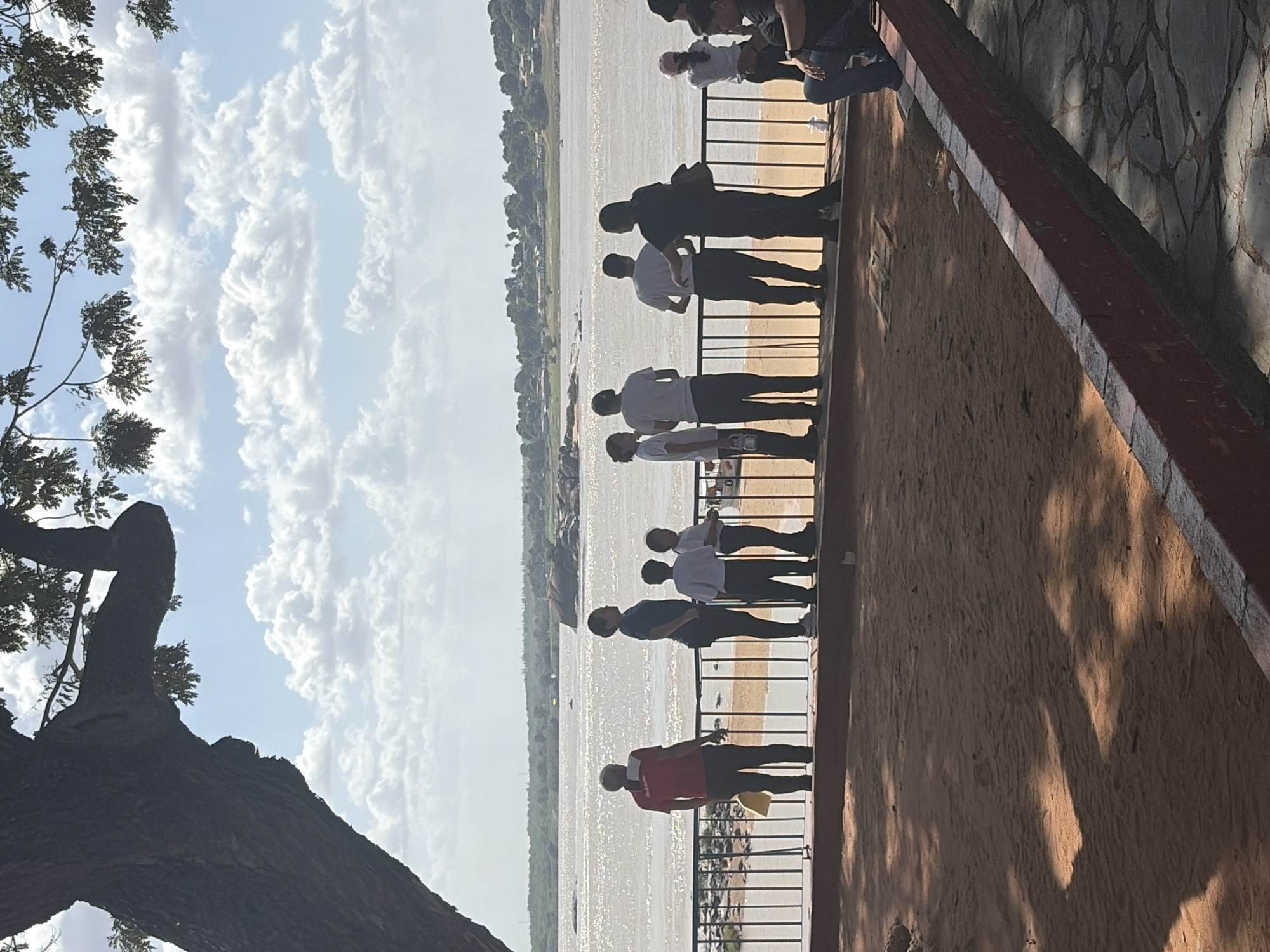 From Ciudad Bolivar, Venezuela, a group of individuals is observed standing along a metal railing, gazing out towards a vast body of water. A line of vegetation can be seen on the opposite shore, and a few boats are in the distance on the water. Fluffy white clouds fill the sky above. The image is taken from a slightly elevated position, with part of a tree branch and trunk visible in the lower left corner. The railing is situated next to a wall with a brick border and a stone patterned section behind it.