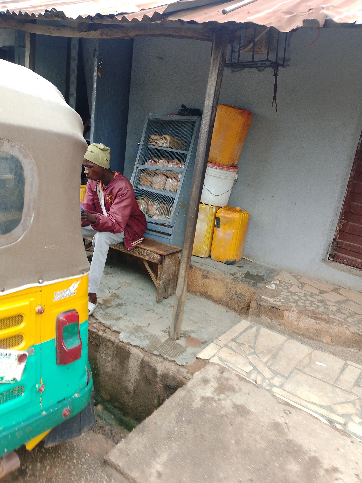In an outdoor, covered area, a young man in a maroon jacket and grey pants sits on a wooden bench. He wears a green beanie. To his left, a portion of a yellow and green auto-rickshaw is visible. Behind the man, a metal shelf unit displays rows of bread loaves in plastic packaging. To the right of the shelf, there are two large yellow plastic containers stacked atop a white bucket, and another yellow plastic container is on the ground. A weathered, corrugated metal roof extends over the scene. The ground is a mix of concrete and cracked tiles, with a visible drainage ditch to the left of the auto-rickshaw. The background is a grey wall, with a brown, slatted window on the right side.