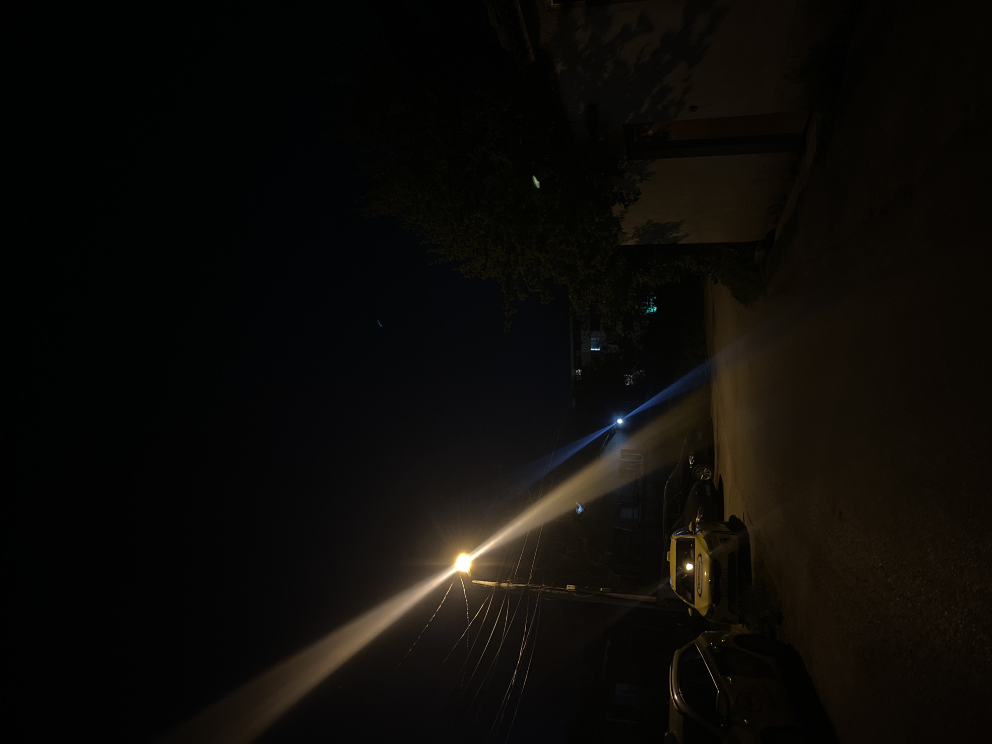 This vertical nighttime photograph captures an urban street or alleyway in Sofia, Bulgaria. The majority of the left side of the image is a dark, unlit sky and ground/roadway. The scene is illuminated by two strong artificial light sources. A prominent, bright yellow-orange streetlight is located in the lower-left portion of the image, casting an intense beam of light diagonally upwards and to the right. Below this light, numerous dark wires or cables extend downwards.
Further up and to the right, a second bluer light source is visible, also emitting a distinct beam. On the right side of the frame, a tall, dark building or wall runs vertically, with shadows of tree branches or leaves visible on its upper part. A tree or dense foliage is present in the upper-middle area, partially obscured by darkness.
Parked against the wall in the lower-right quadrant is a yellow service or utility vehicle, featuring a light bar on its roof. Some dark, unidentifiable text or a logo is visible on its side. Behind this, another dark, partially visible object, possibly another parked vehicle, is present. The overall atmosphere is quiet and still, indicative of a deserted street at night under clear skies. No human activity or discernible text beyond the unidentifiable logo is visible.
