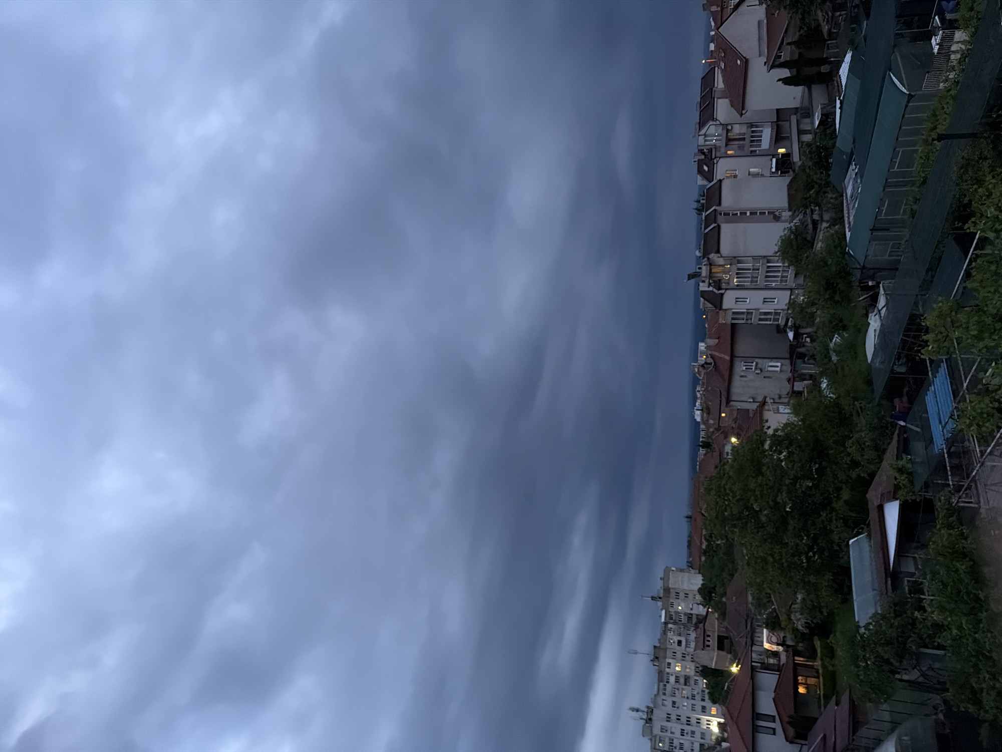 The image shows a cityscape in Burgas, Bulgaria, under a dark, cloudy sky. Buildings with red tile roofs and white facades line the right side of the image, interspersed with trees. Some windows are lit, indicating it is dusk or night. A metal structure, possibly a trellis or fence, runs along the top right. The sky occupies the majority of the frame, dominated by heavy, grey clouds.