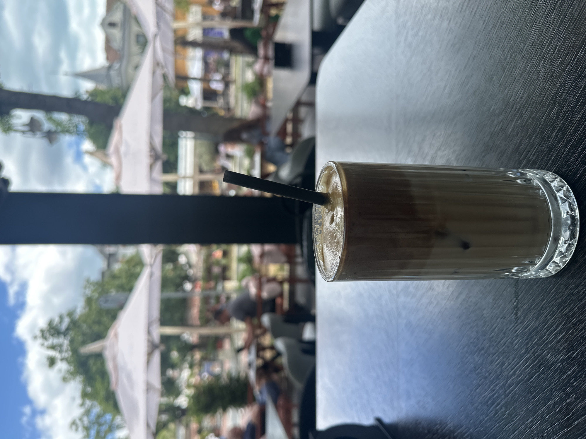 A tall glass of iced coffee with a straw sits on a dark wooden table in the foreground, with an out-of-focus background of an outdoor cafe setting. The cafe features white umbrellas, trees, and people sitting at tables. The sky is visible with clouds. The focus is on the drink, with the background providing context for an outdoor dining experience.