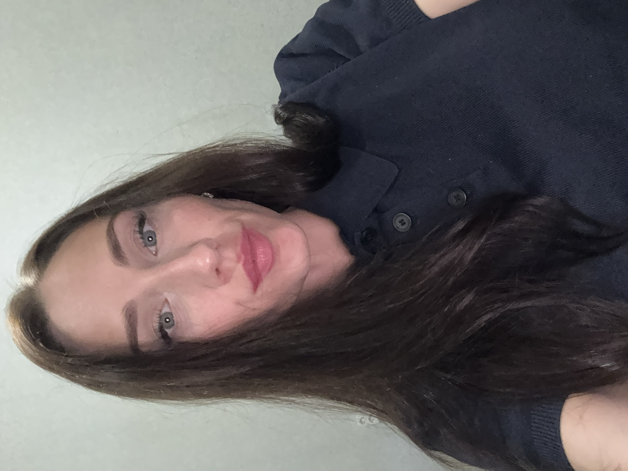 A medium shot captures a woman with long, dark brown hair styled straight, framing her face. She has blue eyes, arched eyebrows, and is wearing pink lipstick. The woman is dressed in a dark blue collared shirt with two buttons. The background appears to be a plain, light-colored wall, suggesting an indoor setting, possibly a residence or office. No specific landmarks or environmental details are visible to indicate a location within Saint Petersburg, Russia.