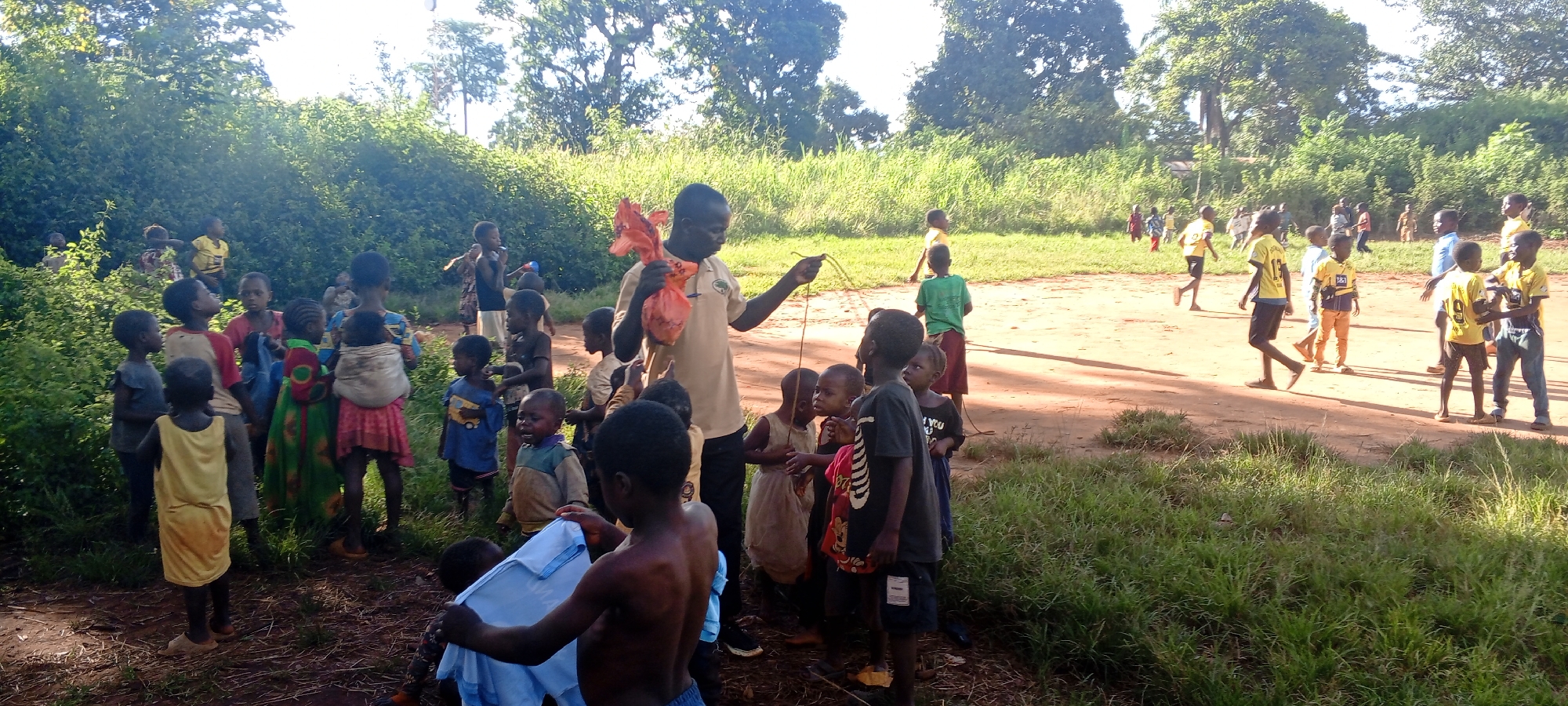 This image depicts an outdoor scene in Ngalula, Democratic Republic of the Congo, characterized by a mix of dense vegetation and cleared ground. The environment consists of lush green bushes and tall grass bordering a reddish-brown dirt field. Mature trees are visible in the background under bright daylight, casting defined shadows.
In the left and central foreground, approximately 15-20 children, ranging from infants carried in wraps to early teenagers, are gathered closely. They wear various casual garments including t-shirts, dresses, and shorts. An adult male stands centrally, wearing a light tan short-sleeved collared shirt with a circular emblem on the left chest. He holds an orange plastic package in his left hand and a thin, brown, flexible object, resembling a rope or string, in his right hand. Many of the children are looking towards this adult. One bare-chested child in the immediate foreground holds a light blue garment.
In the midground and background to the right, approximately 10-15 additional children are visible on the open dirt field. Many of these children wear yellow short-sleeved shirts, some with numbers or logos such as "1881" or "9", suggesting team attire. They are observed moving across the field, some running or walking, indicative of an ongoing or impending activity.